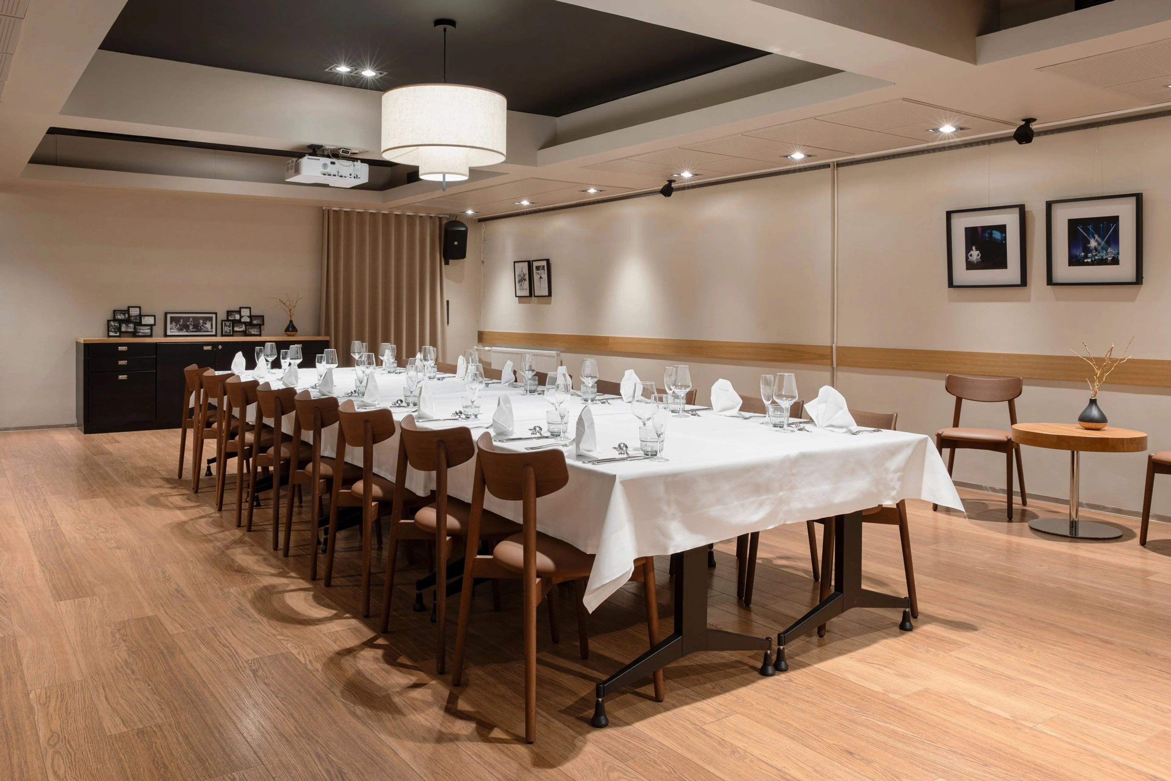 Tyylikäs yksityinen ruokailutila, jossa pitkä pöytä valkoisen pöytäliinan ja kattauksen kera. Puulattia ja lämmin valaistus.