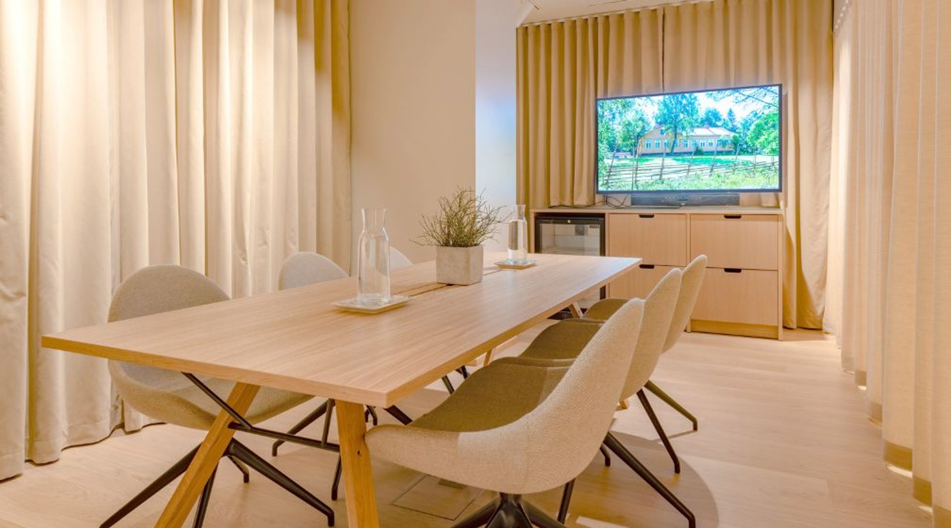 Moderni neuvotteluhuone puupöydällä, valkoisilla tuoleilla ja televisiolla. Tilassa vaaleaa puulattiaa ja pehmeät verhot.