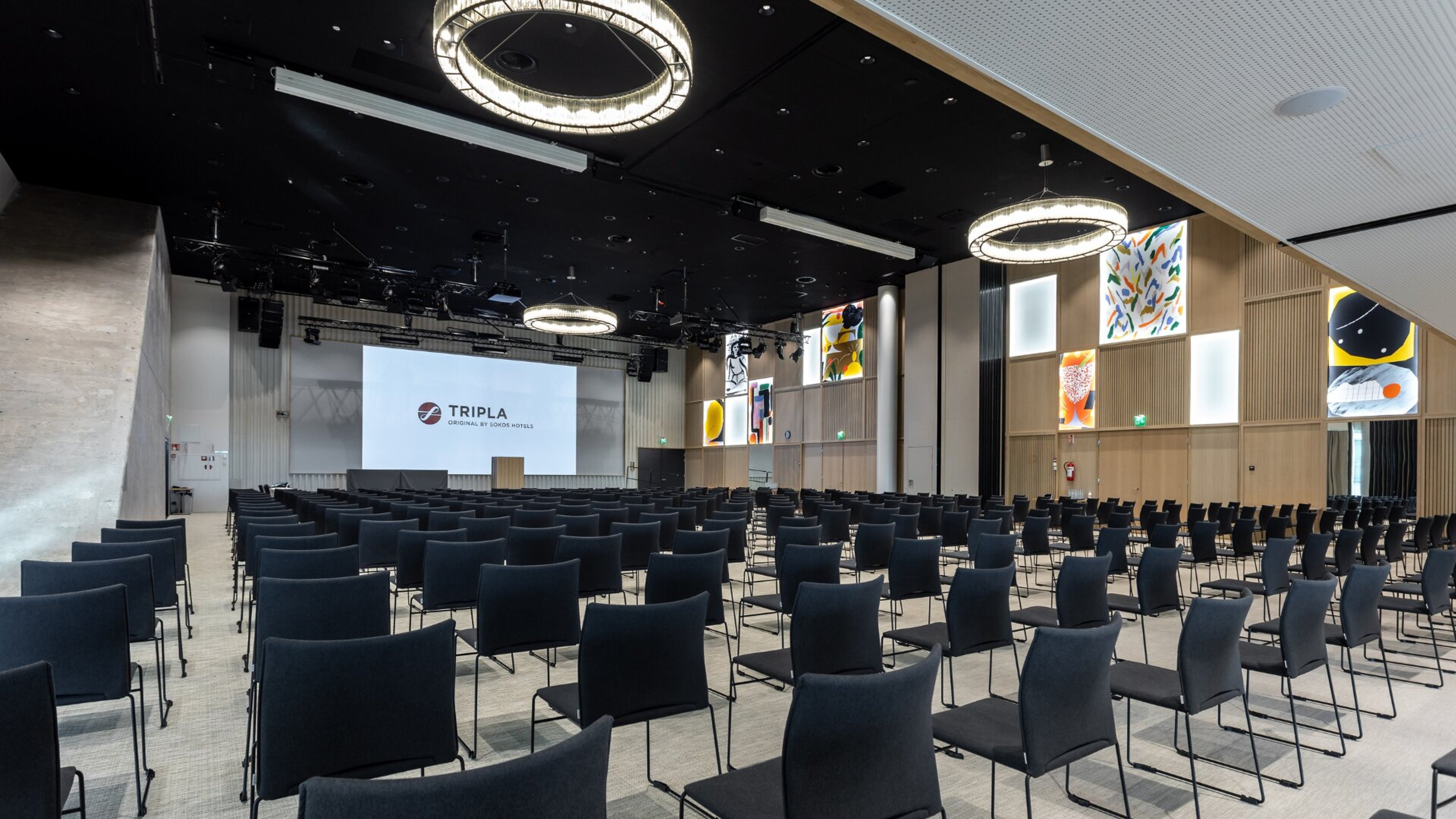 Moderni kokoustilta tummilla tuoleilla, pyöreillä kattovalaisimilla ja värikkäillä taideteoksilla seinillä. Tilava, tasainen lattia ja iso valkokangas edessä.