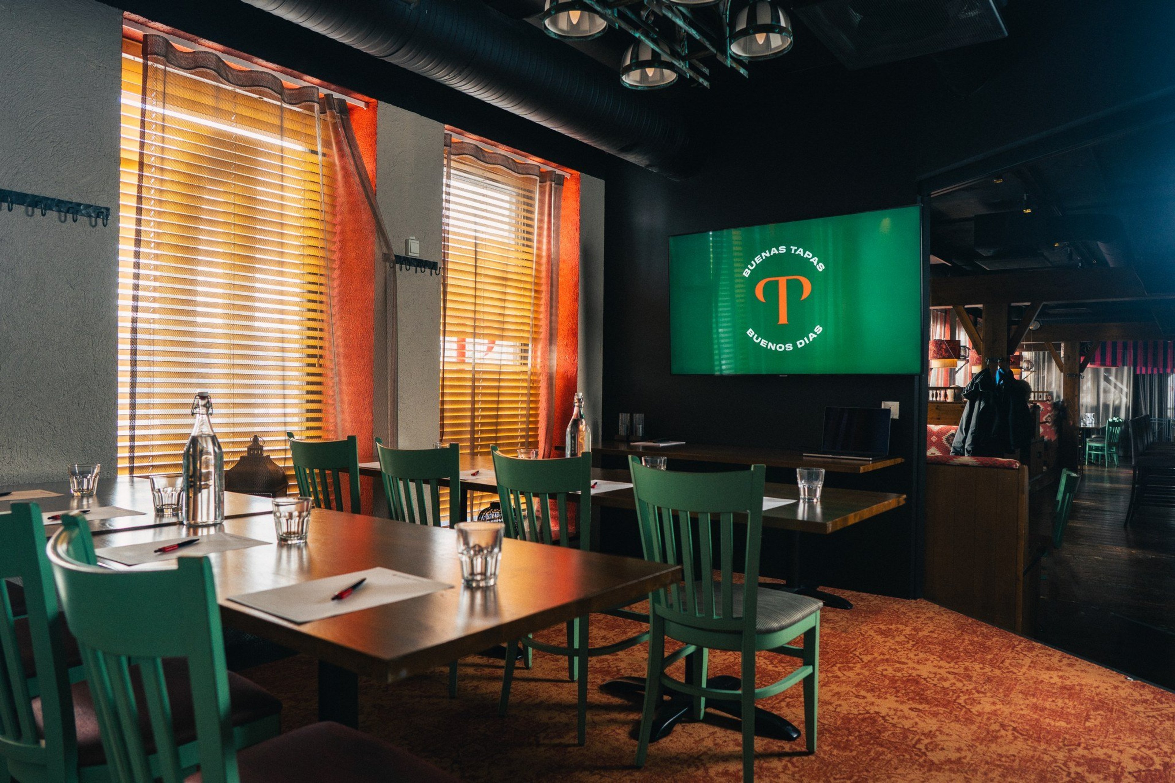 Ravintolan sisätila, jossa vihreitä tuoleja ja puisia pöytiä. Seinällä vihreä näyttö, ikkunoissa sälekaihtimet ja oranssi verhot.