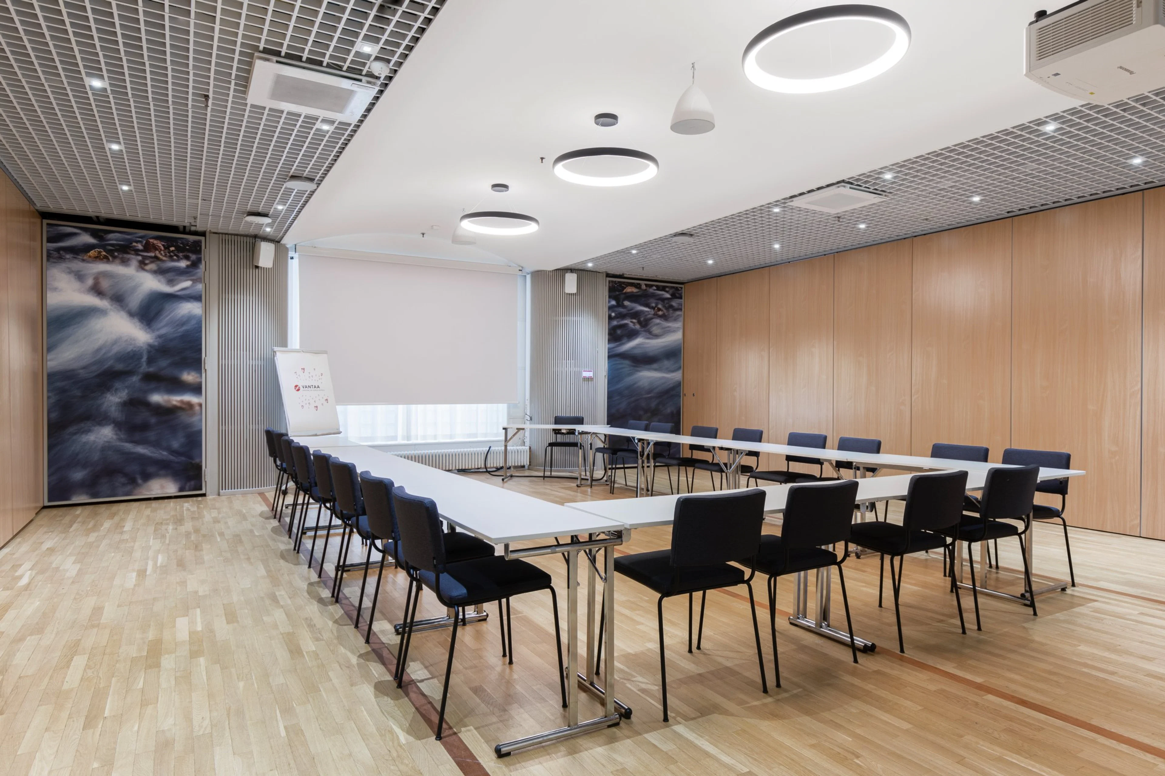 Moderni neuvotteluhuone U-muotoisella pöytäjärjestelyllä. Vaaleat puupaneelit seinillä, tasainen puulattia ja pyöreät kattovalaisimet.