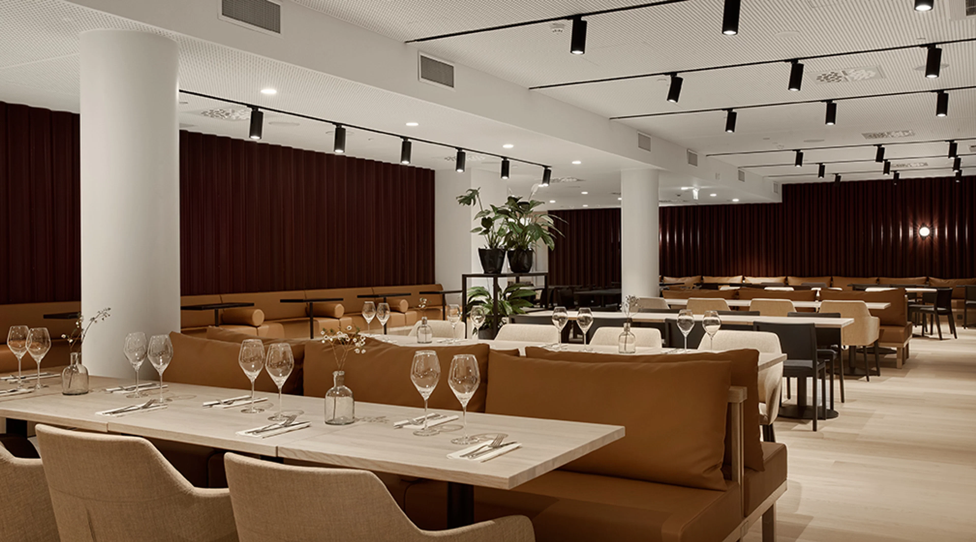 Tyylikäs ravintolasali, jossa vaaleat pöydät, ruskeat sohvat ja tummat puupaneelit seinillä. Avara tila, jossa leveät kulkuväylät.