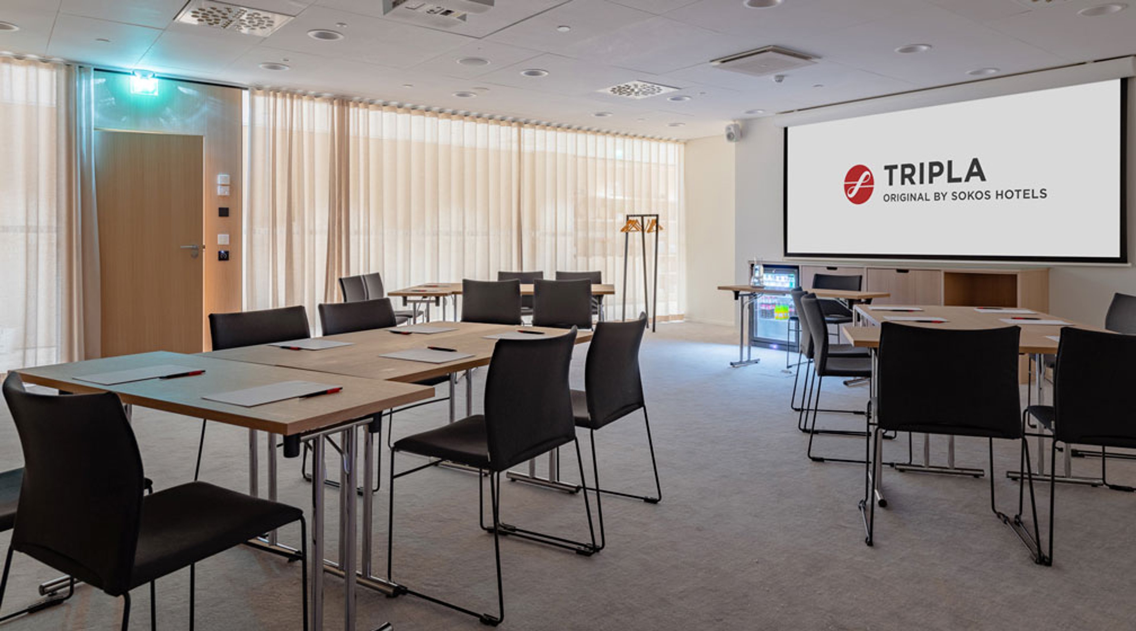 Sokos Hotels Triplan kokoustila, jossa pöytiä ja tuoleja aseteltuna, valkokangas edessä ja beige verhot ikkunoilla.