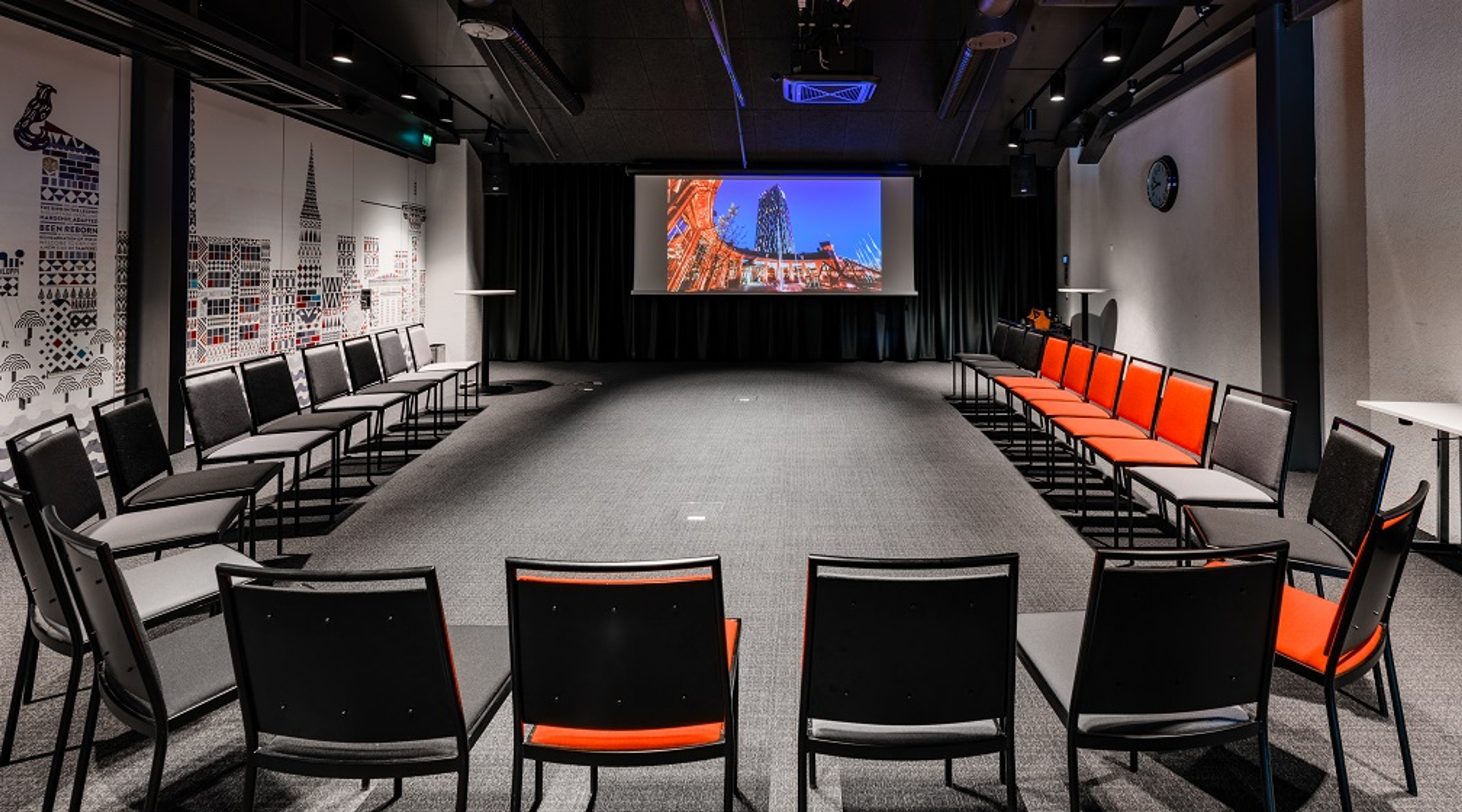 Moderni kokoushuone, jossa tuolit on järjestetty U-muotoon. Oranssi-harmaita istuimia, projektorinäyttö edessä ja tyylikäs citykaupunkikuviointi seinällä.