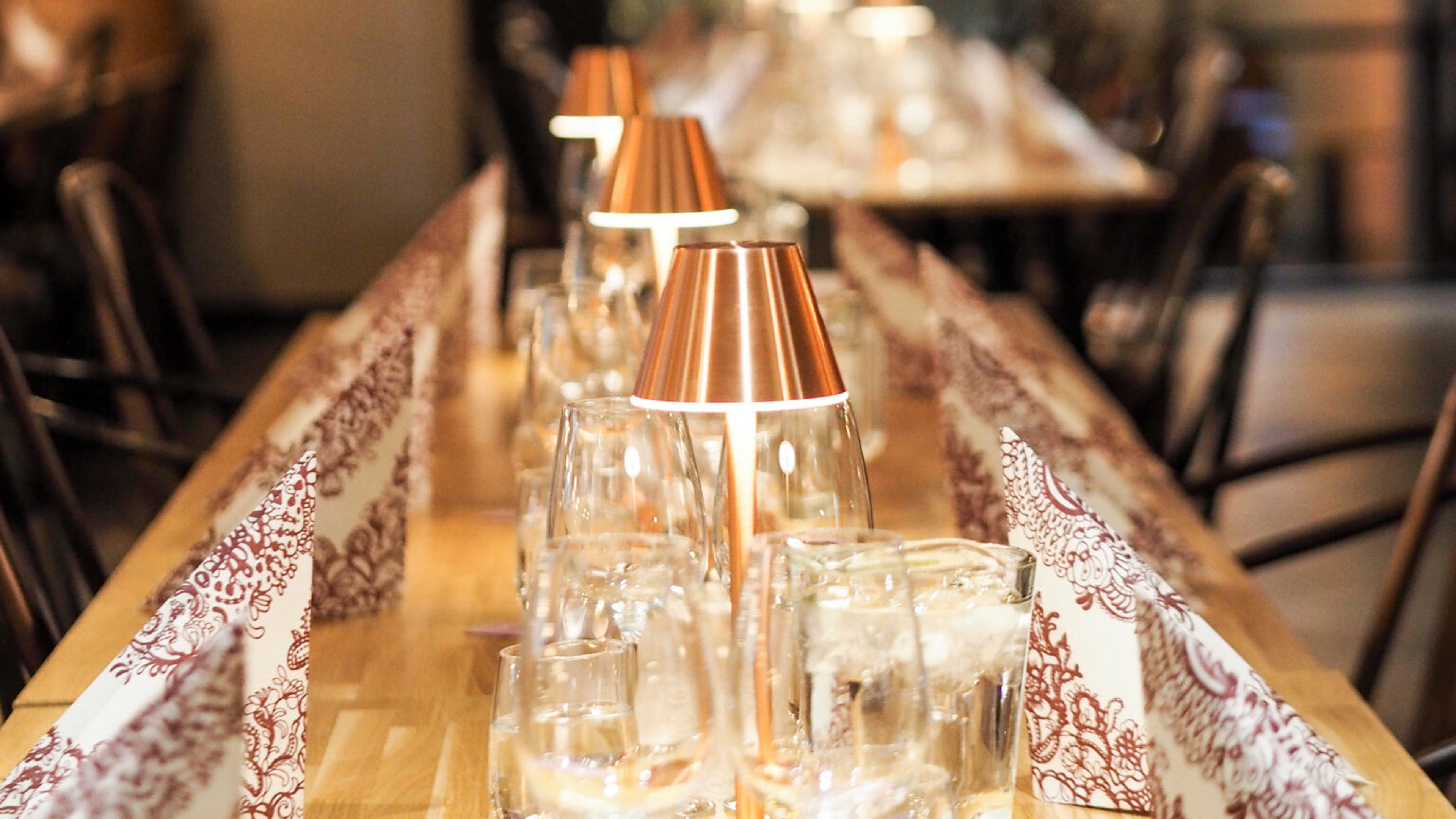 Pitkä puinen pöytä koristelluilla pöytäliinoilla, joissa kuparinvärisiä pöytävalaisimia ja laseja. Tummat tuolit reunustavat pöytää.