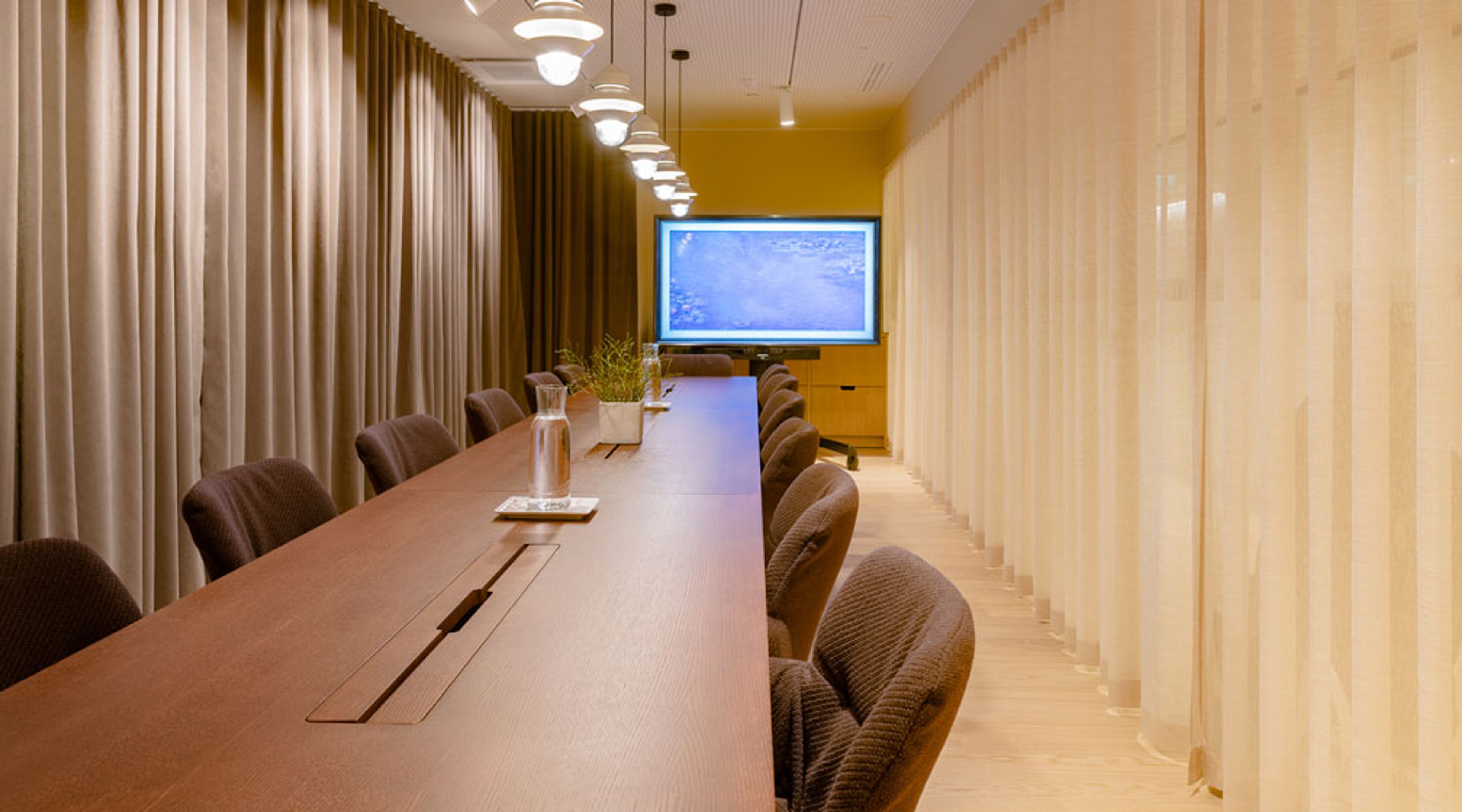 Moderni kokoushuone, jossa pitkä puinen pöytä, harmaat tuolit, valkoisia seiniä ja keltainen takaseinä näytöllä. Katosta riippuvat valaisimet.