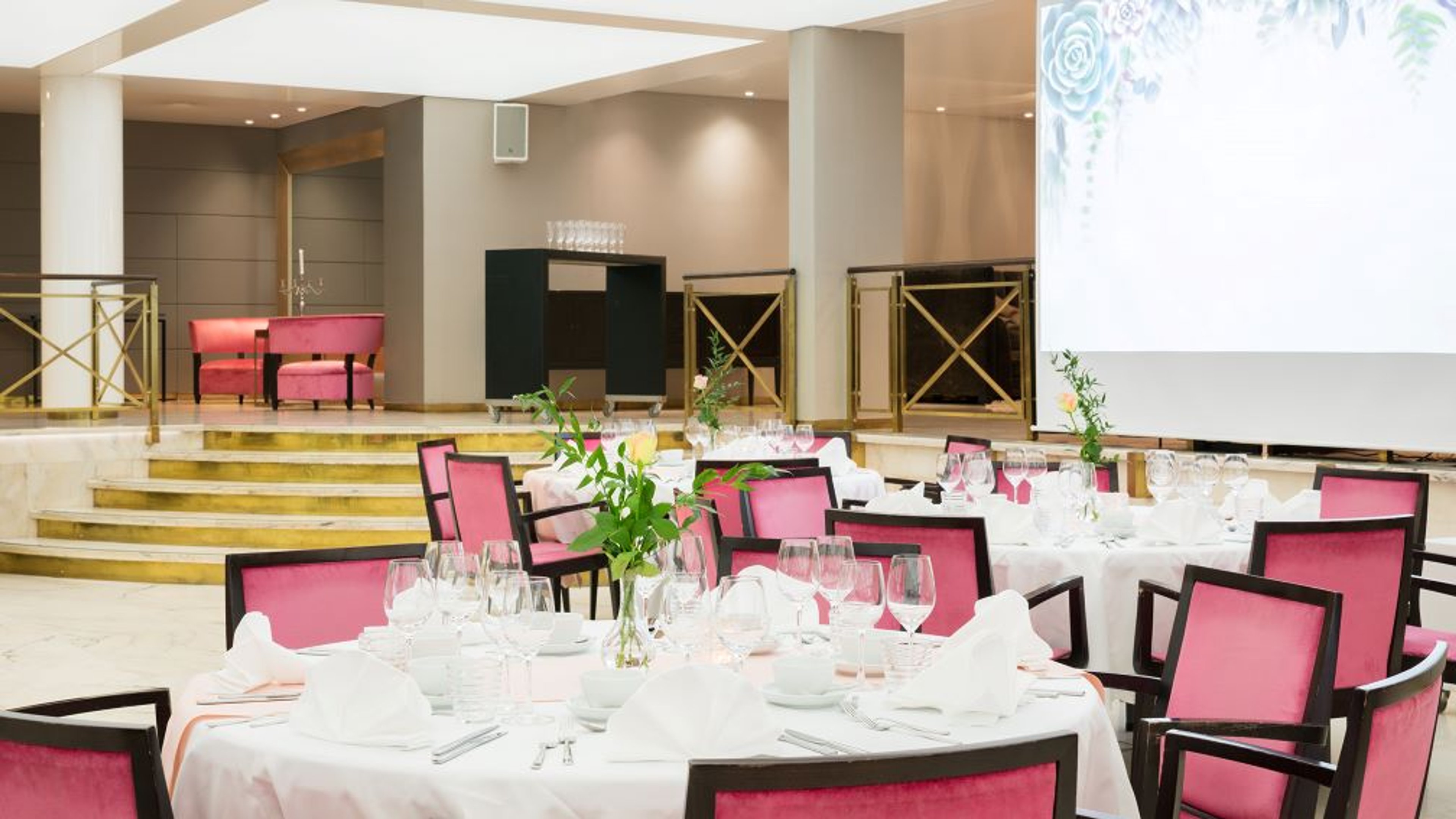 Juhlasali, jossa elegantit valkoiset pöydät ja pinkit tuolit. Taustalla korotettu alue ja valkokangas. Portaat nousevat lavalle.