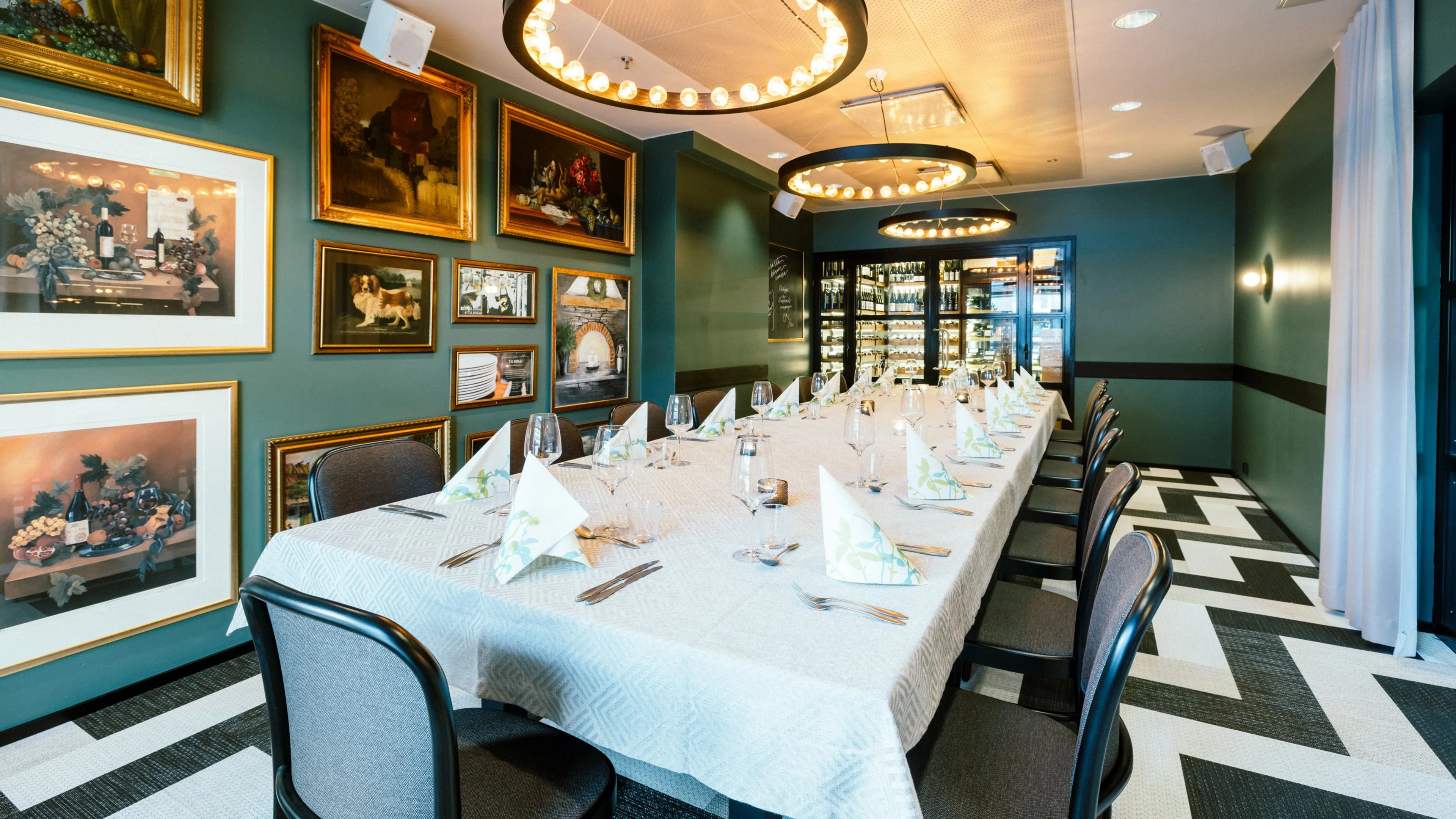 Tyylikäs yksityinen ruokailutila vihreillä seinillä, taide kehyksissä, kolme rengasvalaisinta, pitkä pöytä valkoisella liinalla ja lasi-viinikaappi taustalla. Geometrinen lattiakuvio.