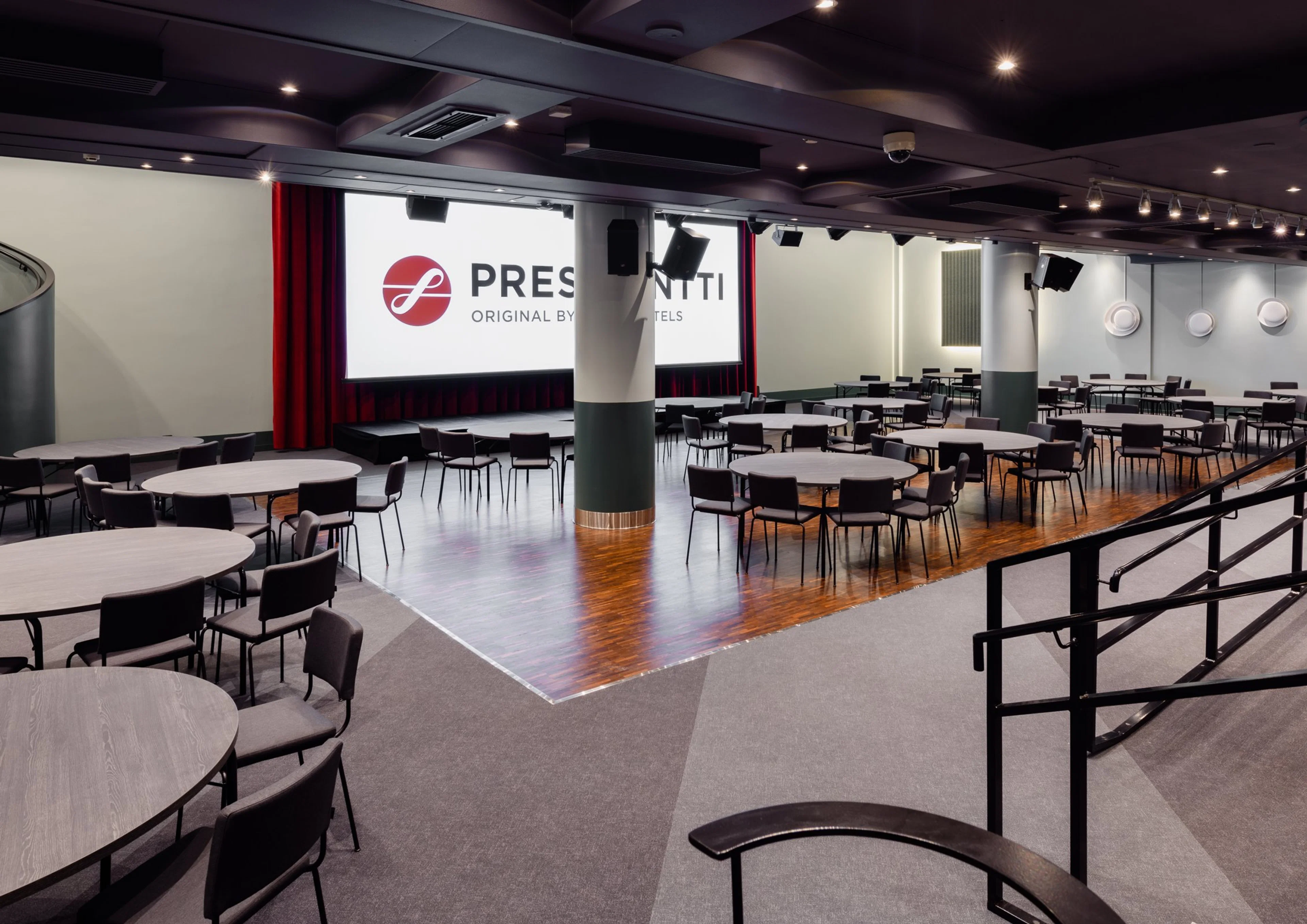 Moderni auditorio pyöreillä pöydillä ja tuoleilla, puulattia keskellä, valkokangas ja punaiset verhot edessä, harmaa kokolattiamatto reunoilla.