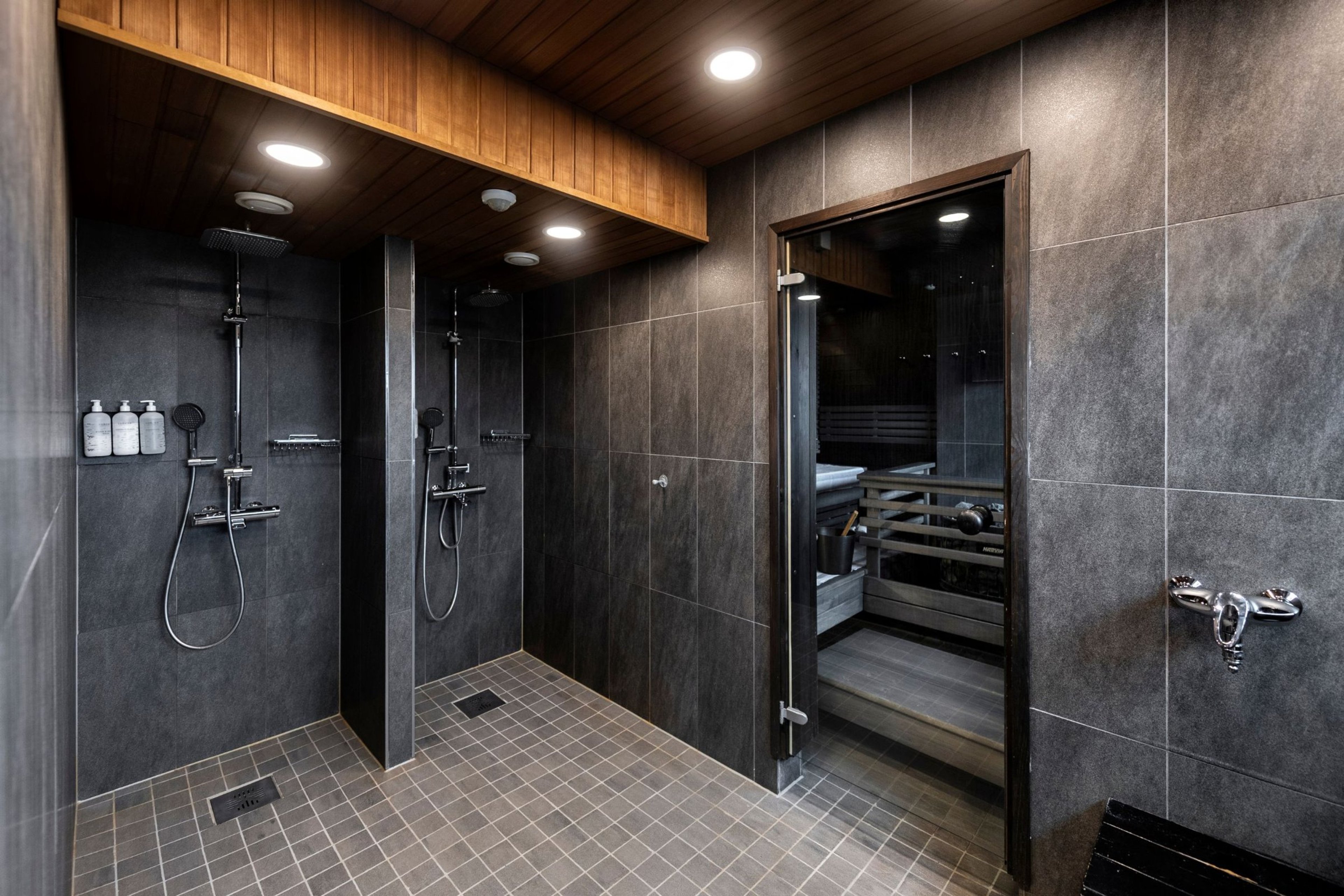 Moderni kylpyhuone, jossa kaksi avointa suihkua ja lasiseinäinen saunan sisäänkäynti. Tummanharmaat laatat ja puinen katto luovat tyylikkään ilmeen.