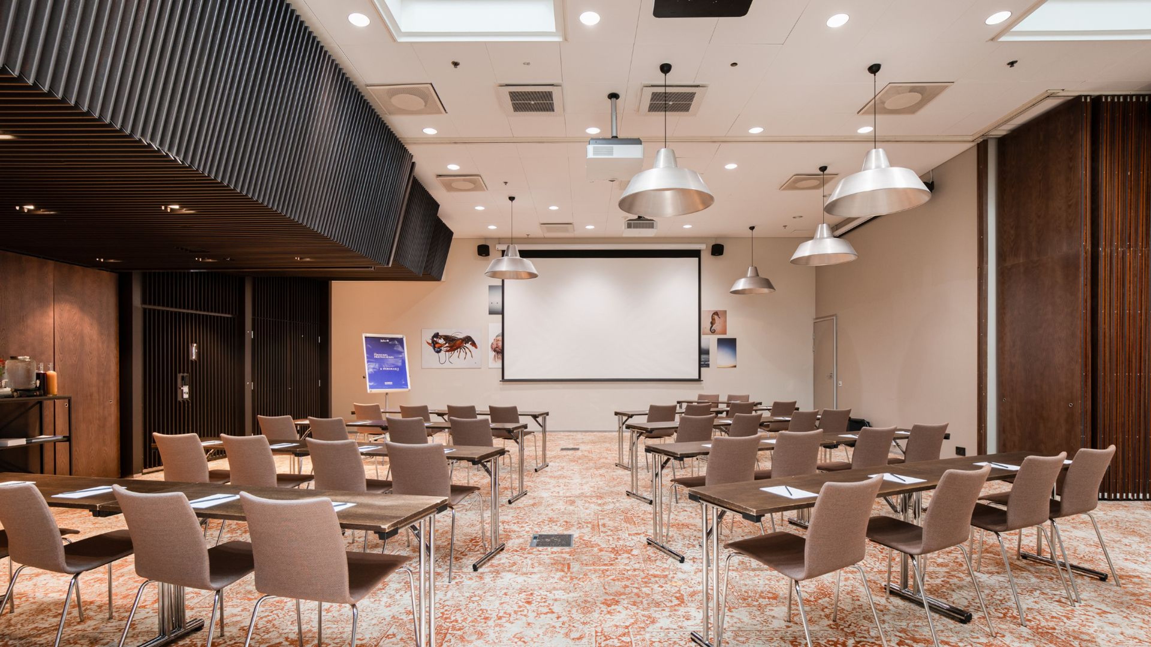 Moderni kokoushuone tuoliriveillä ja valkokankaalla. Tilassa tasainen lattiapinta, hyvä valaistus ja leveät kulkureitit.