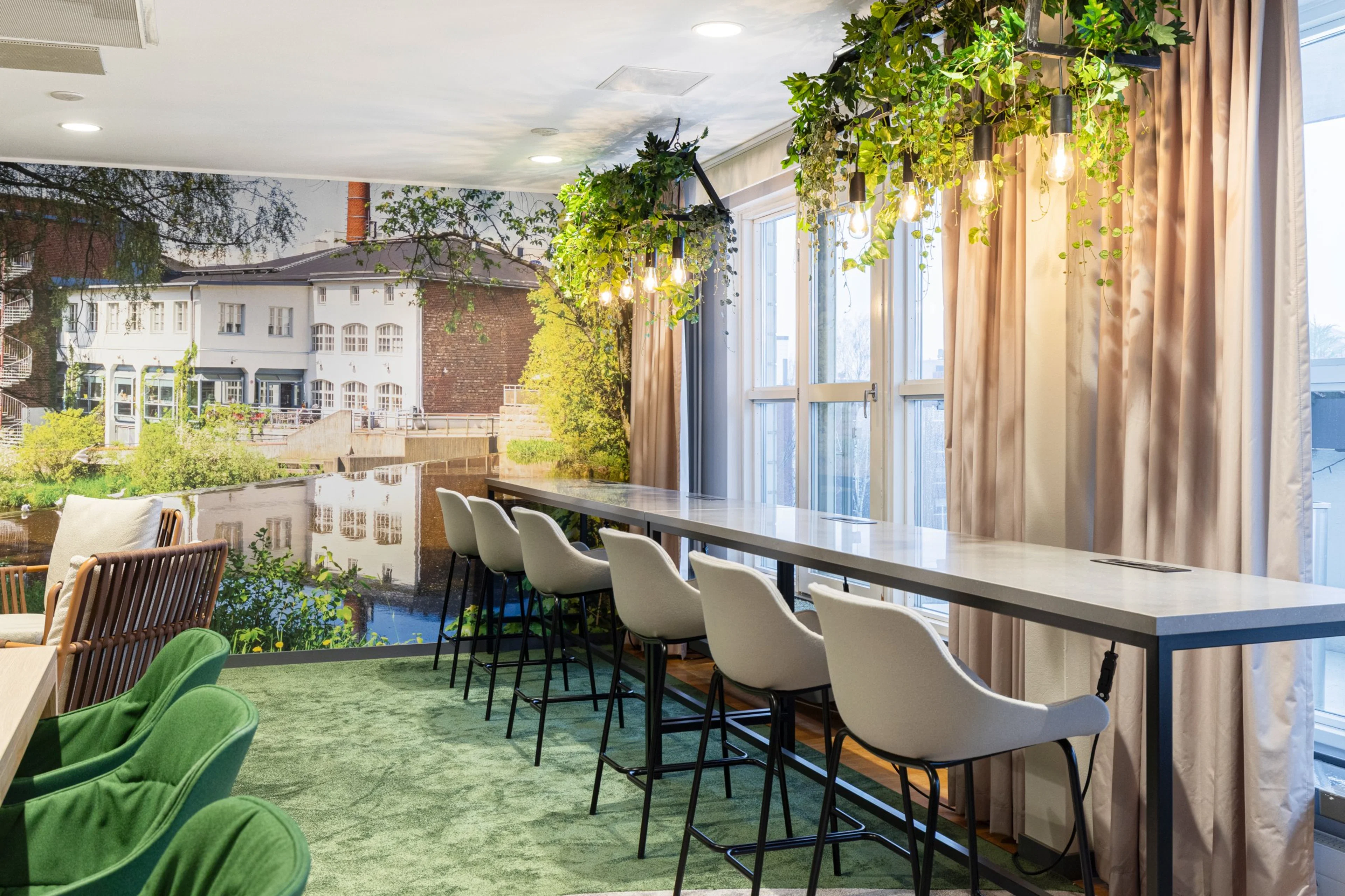 Vihreä loungetila, jossa pitkä baaripöytä valkoisilla tuoleilla, riippuvia viherkasveja, ja ikkunasta näkyy vanha rakennus vedellä.