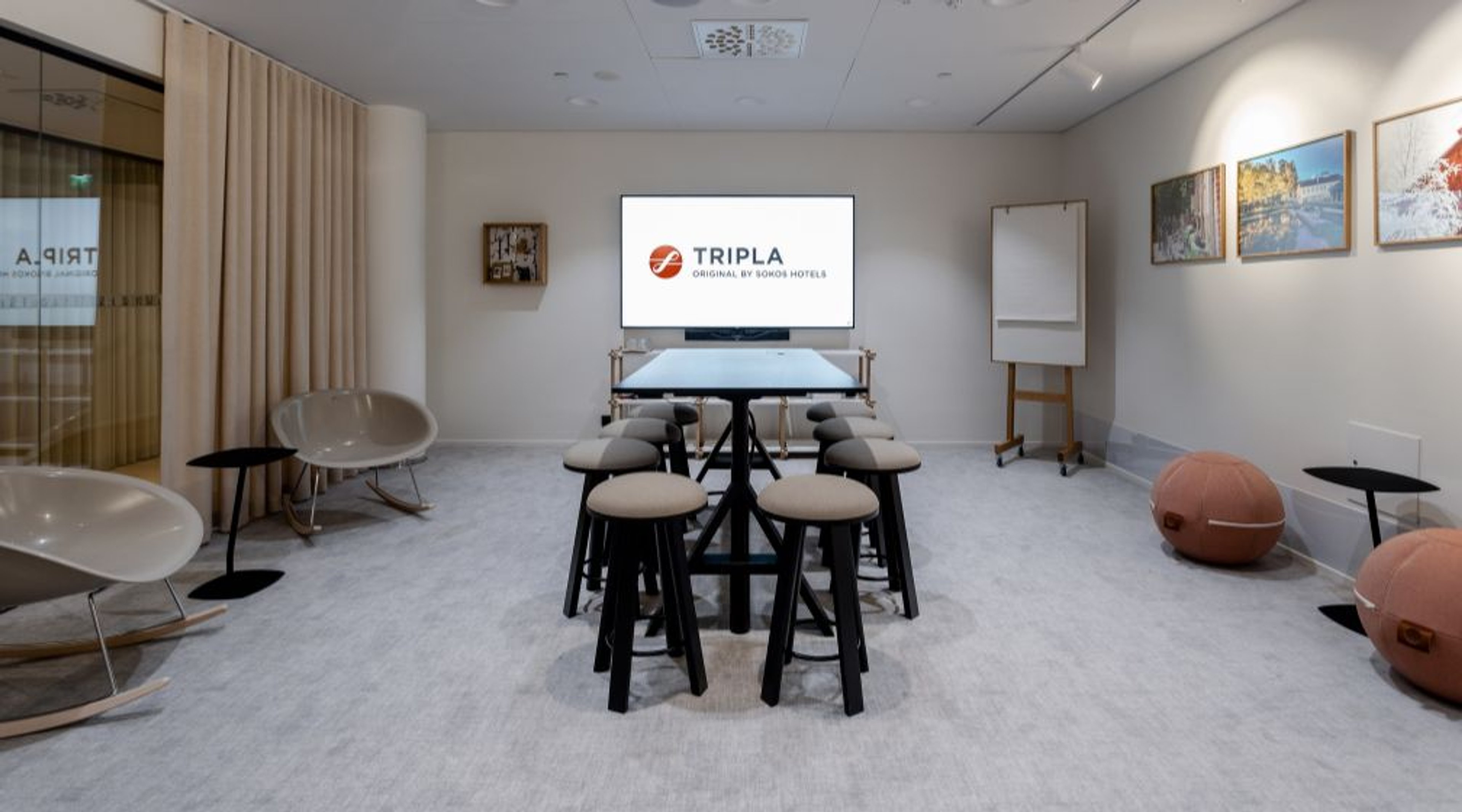 Moderni neuvotteluhuone vaalealla lattiamatolla, jossa on korkea pöytä ja baarituolit, valkotaulu ja näyttö. Seinillä on taidetta ja sivuilla mukavia istuimia.