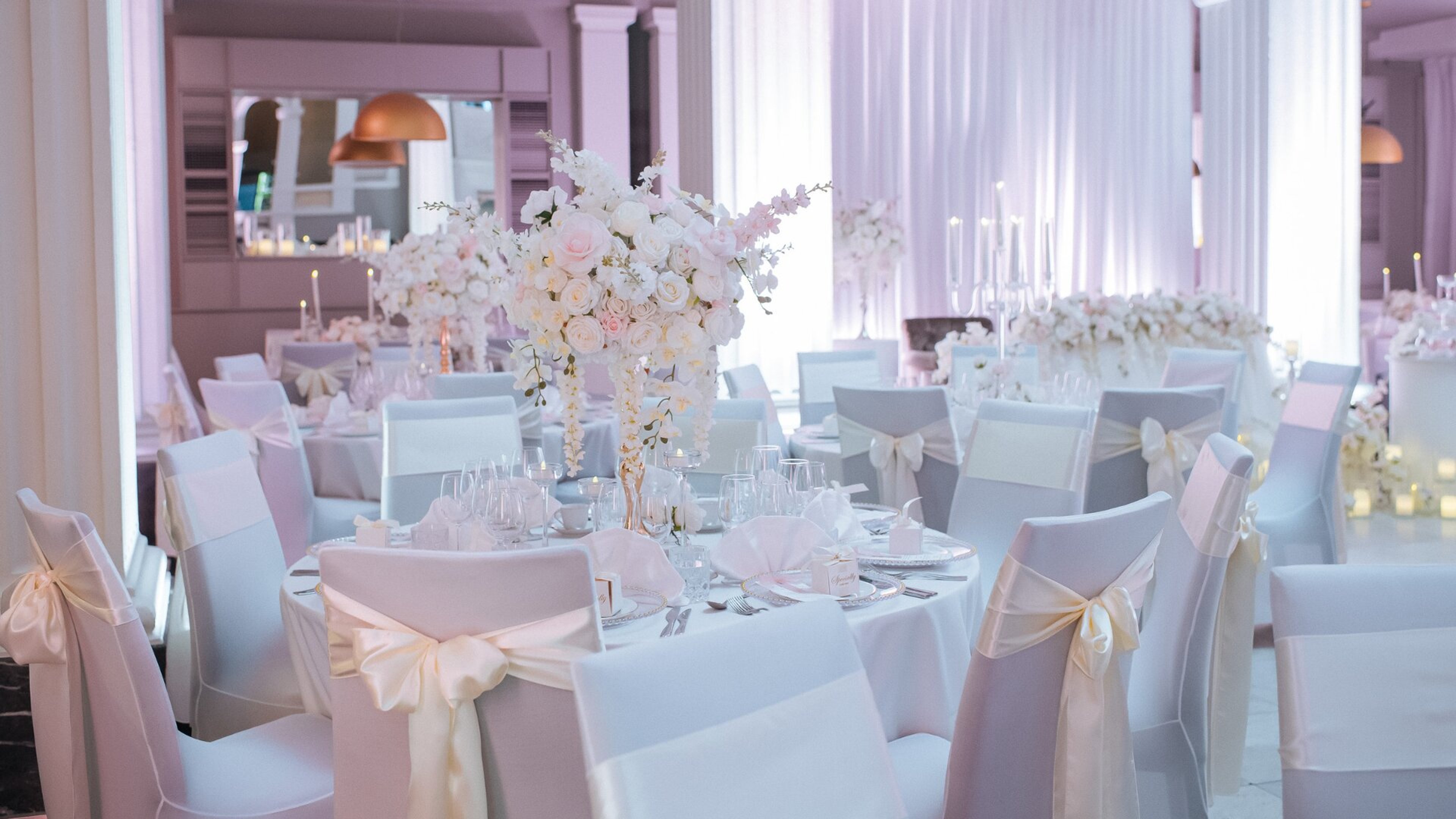 Juhlatila, jossa valkoisia pöytiä ja tuoleja valkoisin rusetin koristeluin. Keskellä pöytää korkea kukka-asetelma vaaleanpunaisilla ja valkoisilla kukilla.