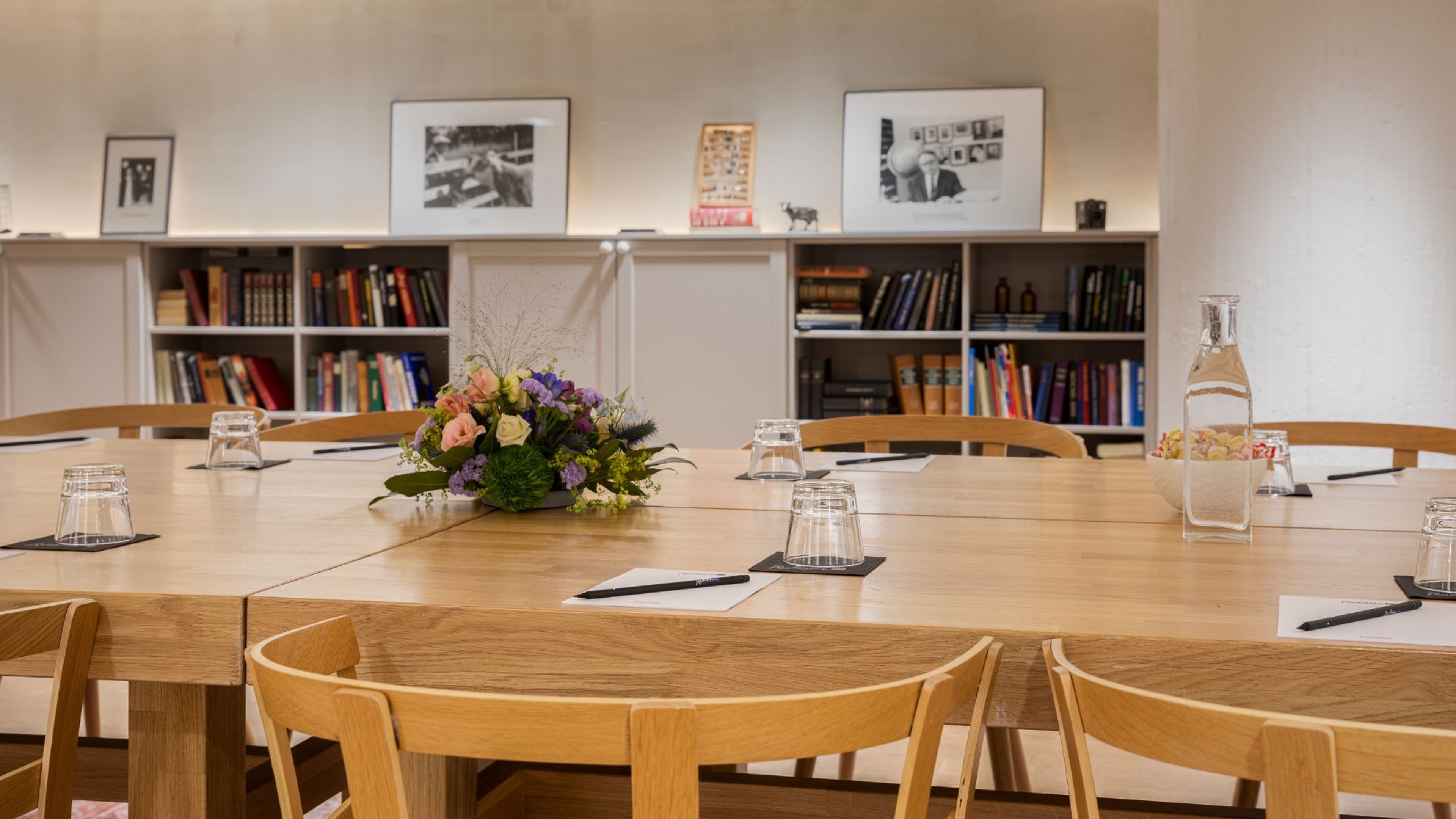 Kokoushuone puisella pöydällä, jonka keskellä kukka-asetelma. Pöydällä vesilaseja ja papereita. Seinustalla kirjahyllyjä ja kehystettyjä valokuvia.