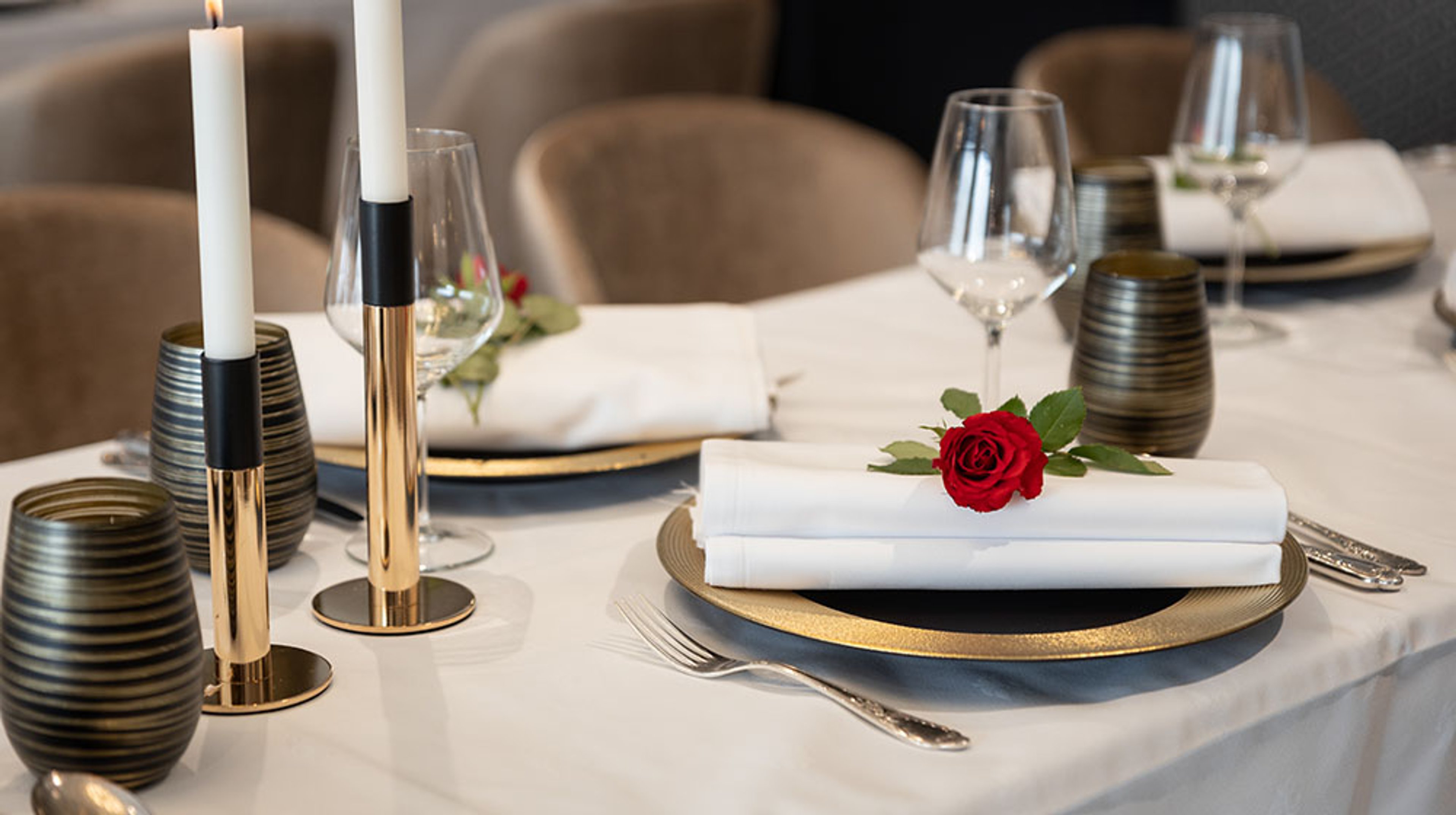 Elegantisti katettu ravintolan pöytä, jossa valkoinen liina, kultaset lautaset ja kynttilänjalat sekä valkoinen servetti punaisen ruusun kanssa.