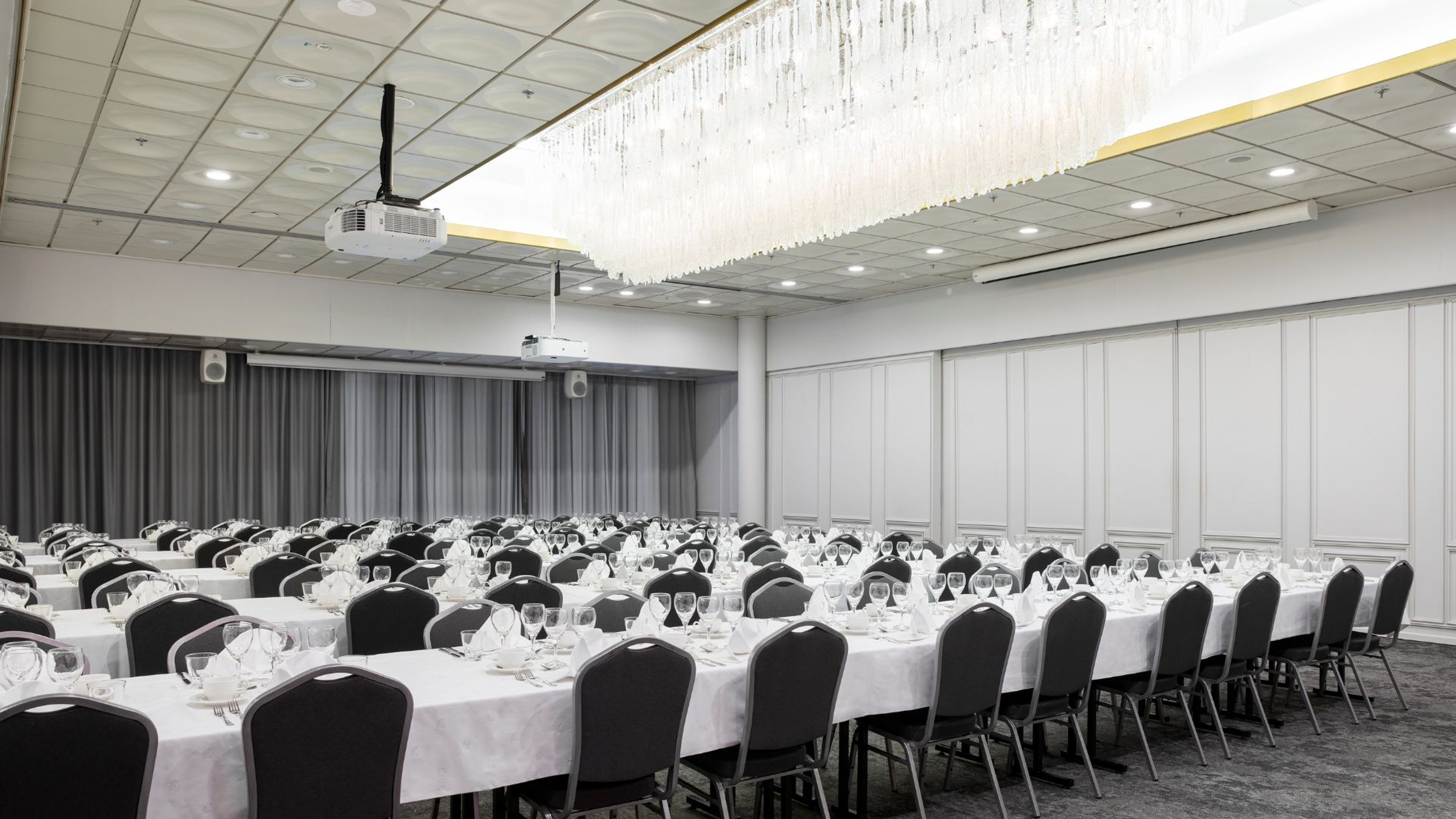 Juhlatila, jossa pitkät pöydät katettuina valkoisin pöytäliinoin ja ruokailuvälinein. Kristallikruunu katossa, seinillä harmaat verhot ja valkoiset paneelit.