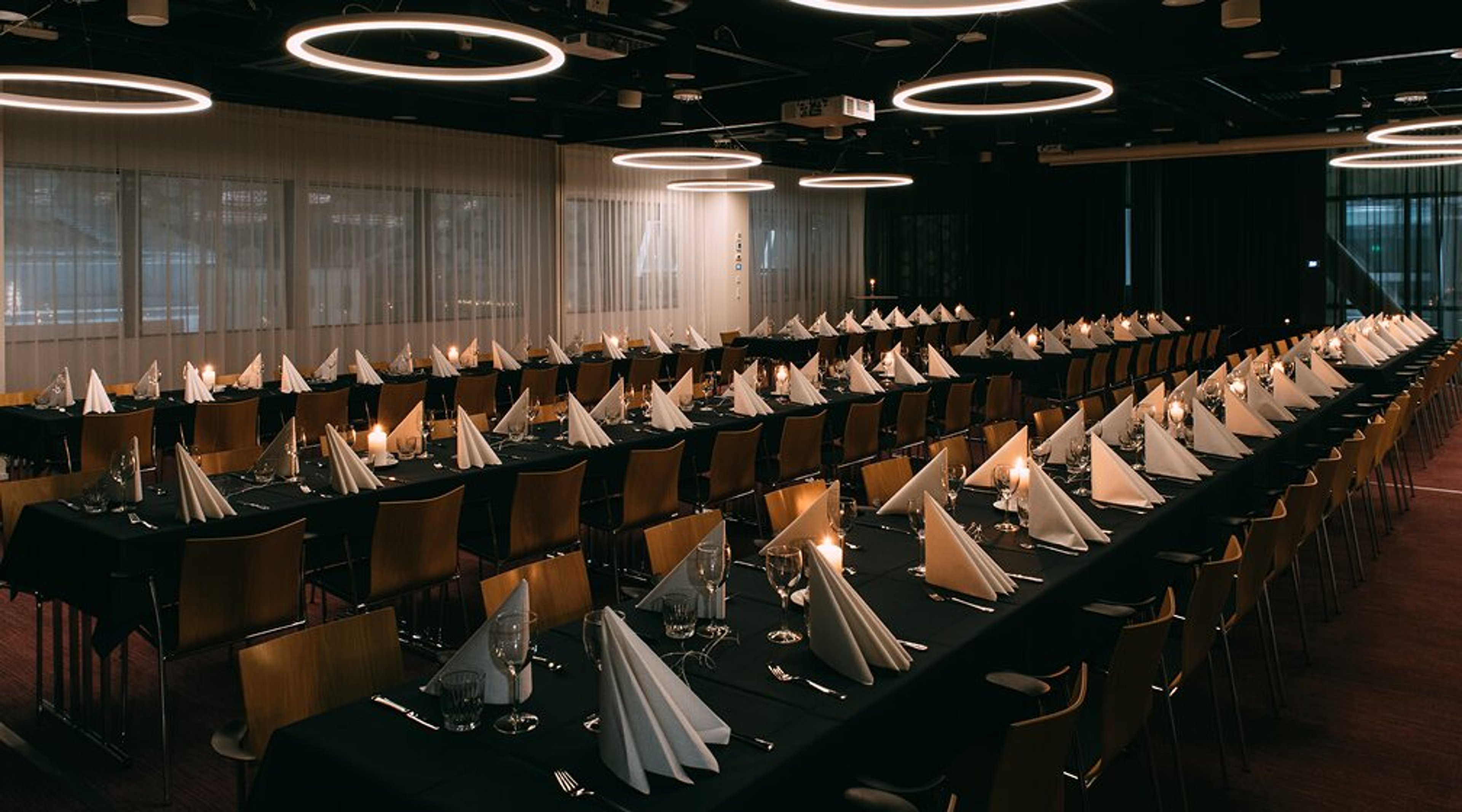 Pidulik söögikorter pikkade laudadega, kaetud mustade laudlinadega, volditud valged salvrätikud ja lauanõud. Laes rõngakujulised valgustid.