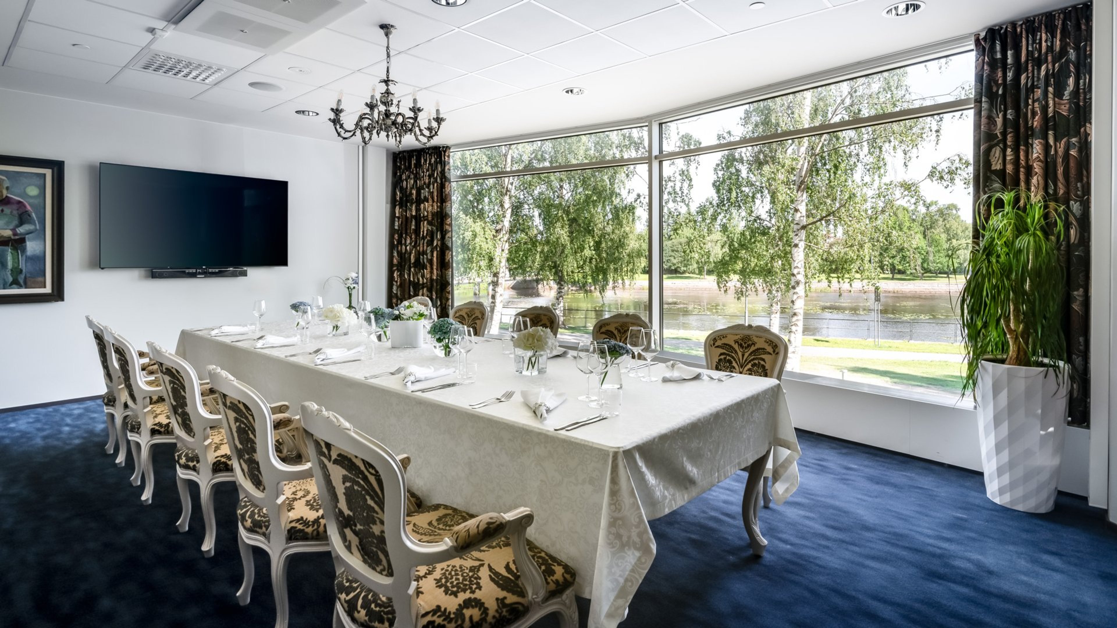 Elegantisti katettu kokoushuone, jossa pitkä valkoinen pöytä, koristelliset tuolit, sininen lattia ja suuret ikkunat joesta ja puista.