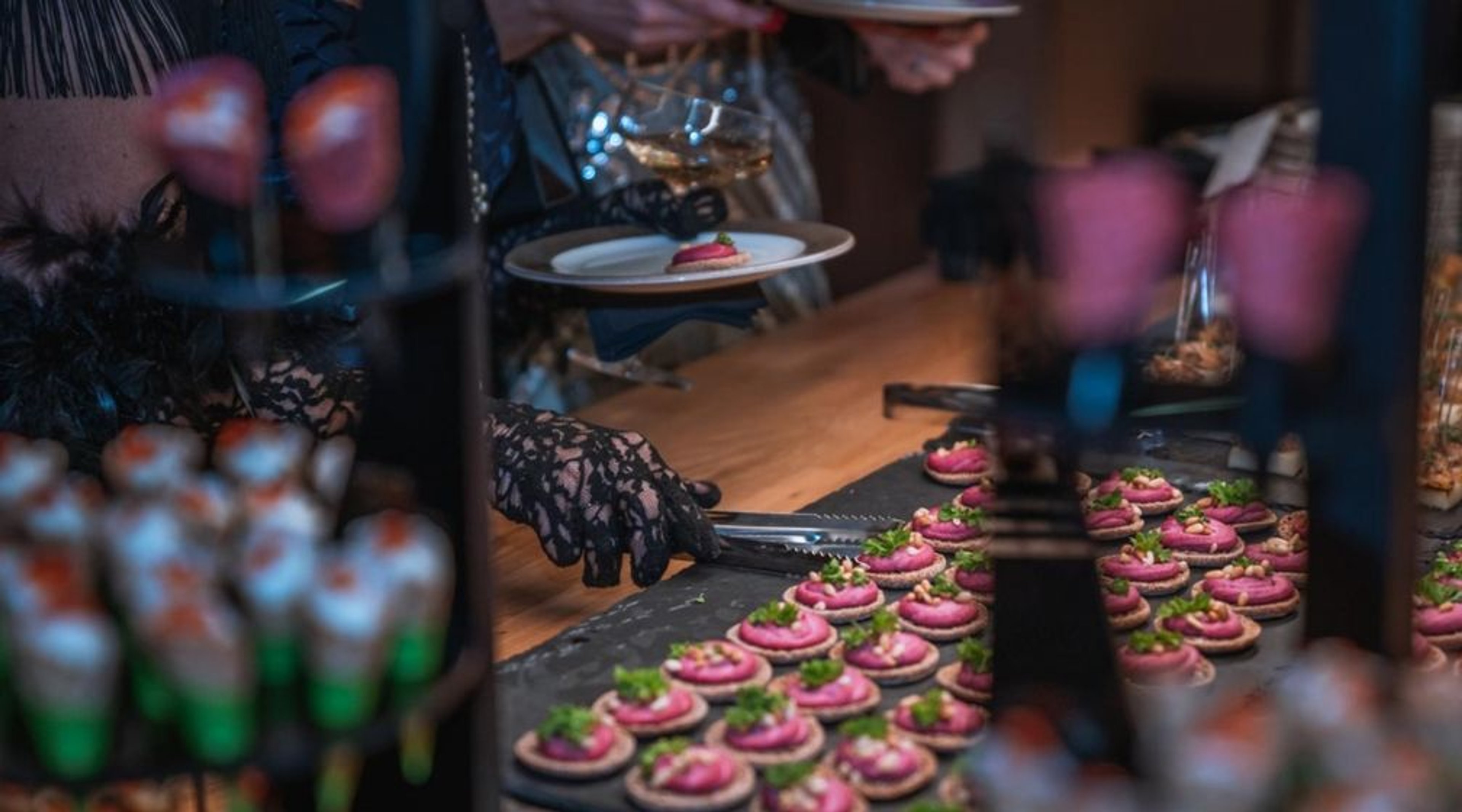 Tarjoilupöytä, jossa käsineilliset kädet ottavat cocktailtarjottimelta pieniä pinkkejä voileipäkakkuja vihreällä koristeella.