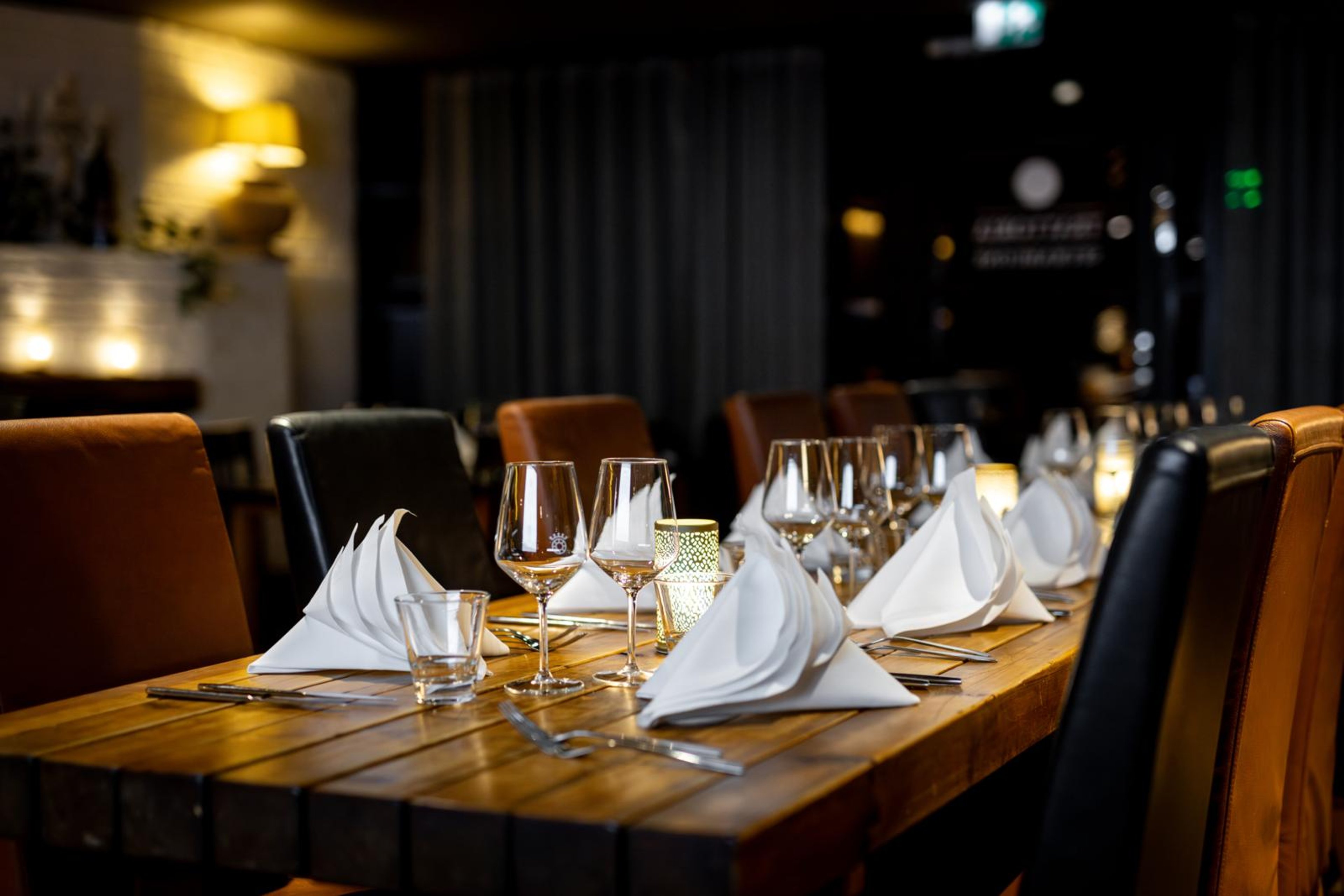 Elegantisti katettu puupöytä ravintolassa, jossa on taitetut valkoiset servietit, viinilasit ja kynttilät. Tuolit ovat ruskeaa ja mustaa nahkaa.