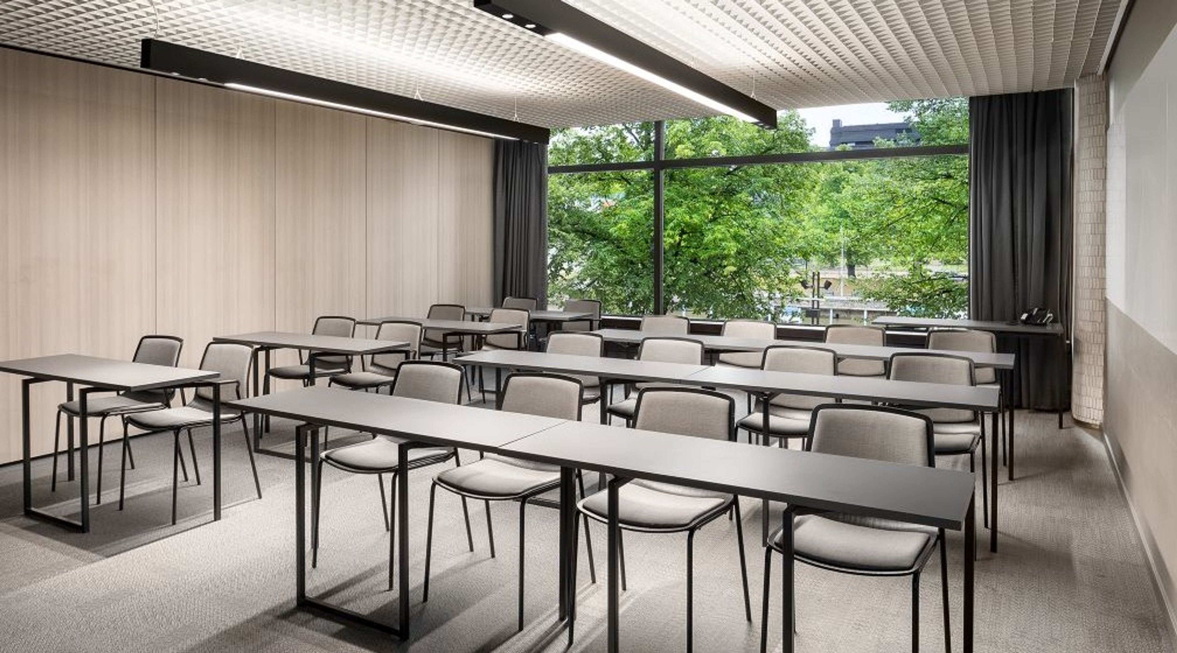 Modern meeting room with long tables and chairs arranged in rows. Large windows overlook green trees. Room features flat carpeting and good lighting.