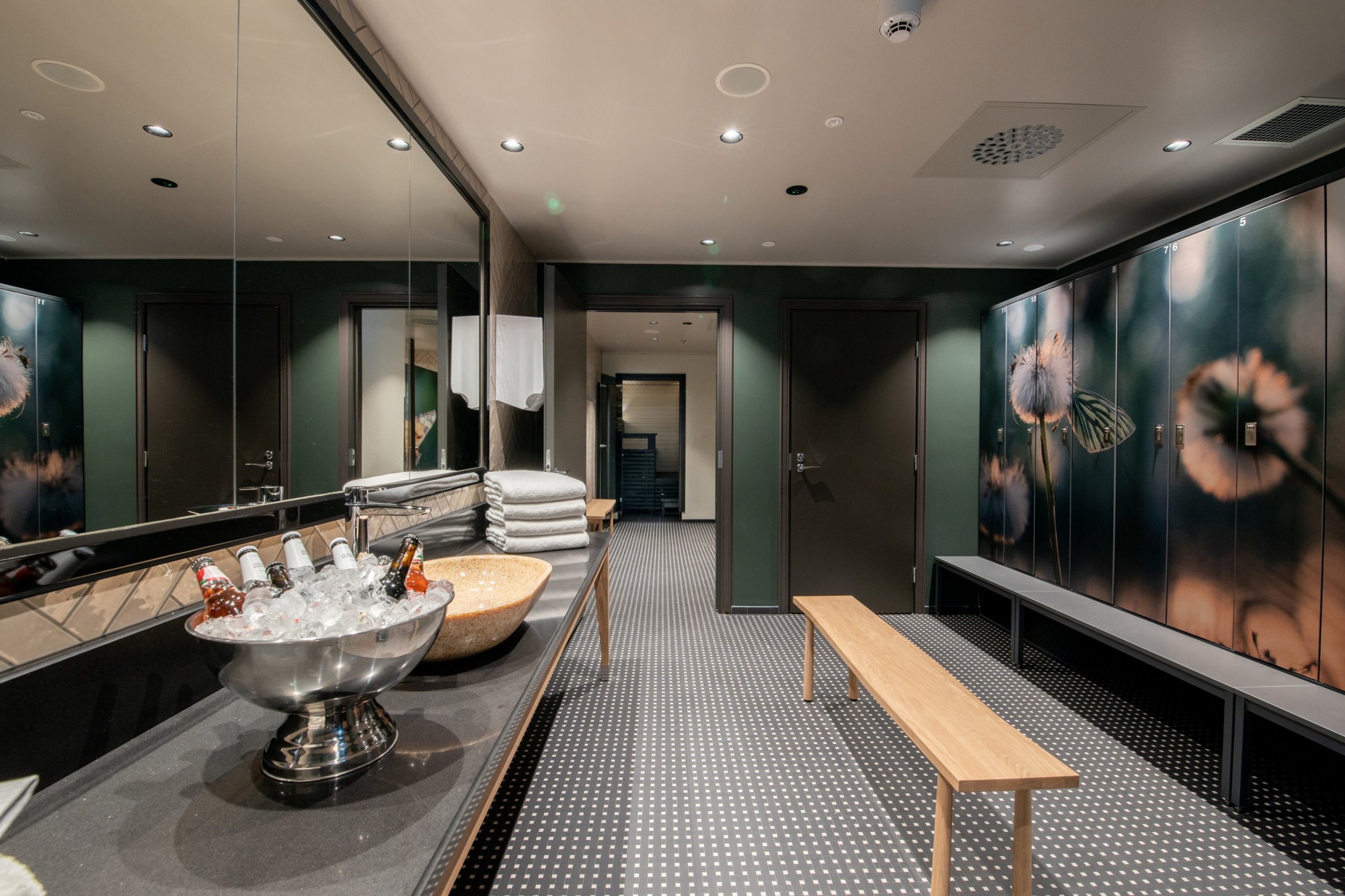 Moderni saunan pukuhuone, jossa vihreitä seiniä, puinen penkki ja kukkakuvioisia kaappeja. Tasolla on pyyhkeitä ja viilentymässä juomia.