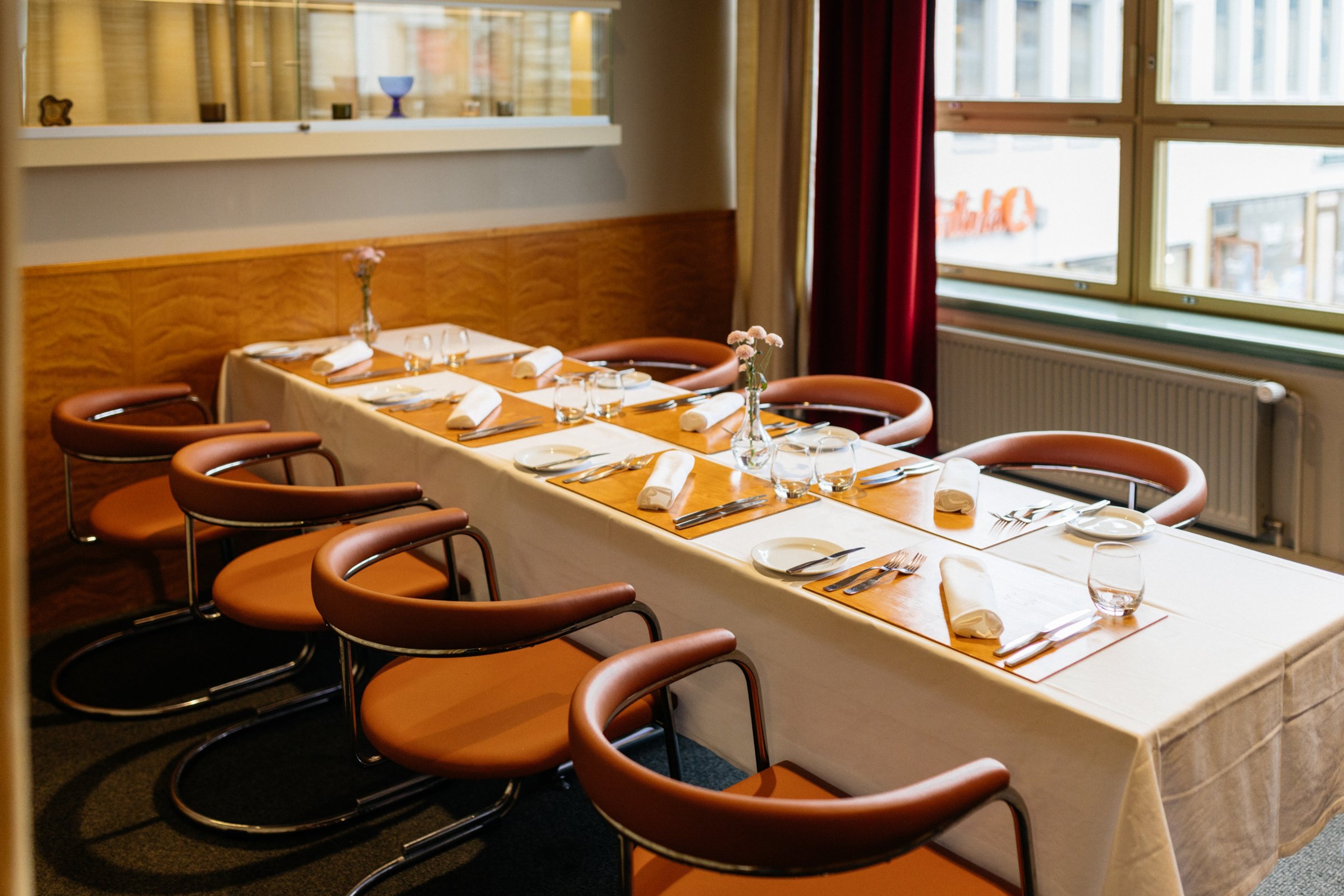 Långt bord i restaurang, dukat för åtta personer. Runt bordet orangea runda stolar, träpaneler på väggarna och fönster med röda gardiner.