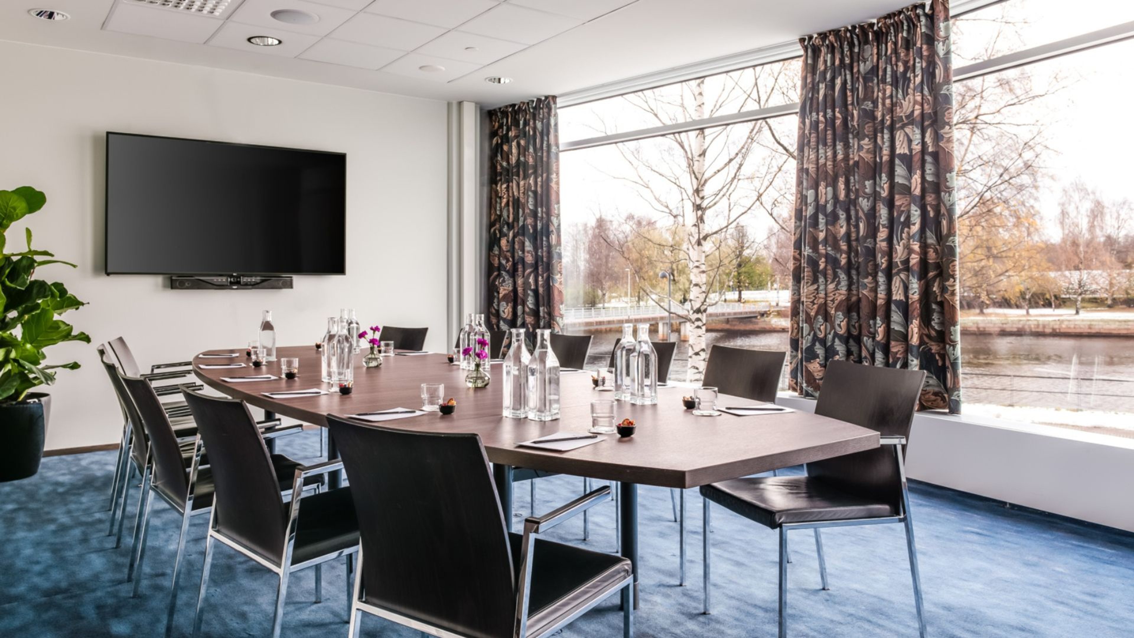 Moderni neuvotteluhuone, jossa on puinen pöytä, mustia tuoleja, suuri televisio seinällä ja lattiasta kattoon ulottuvat ikkunat jokimaisemalla.