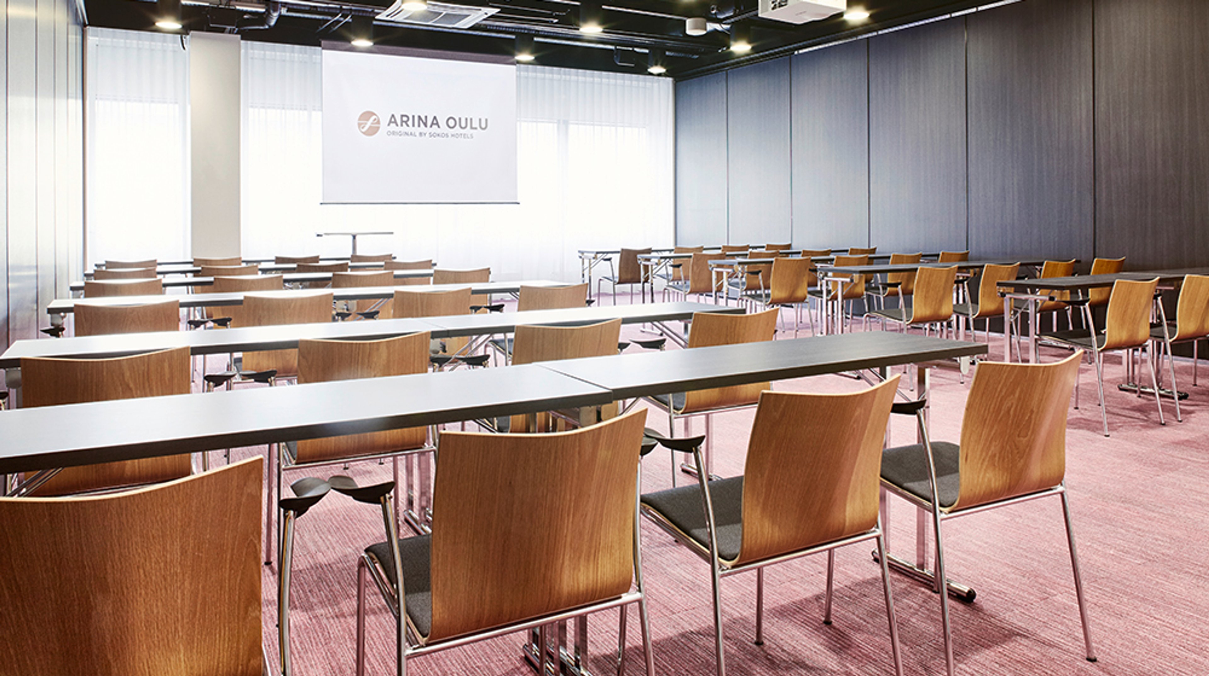 Kokoustila, jossa puiset tuolit ja valkoiset pöydät rivissä. Punainen kokolattiamatto ja valkokangas edessä. Tila on hyvin valaistu.