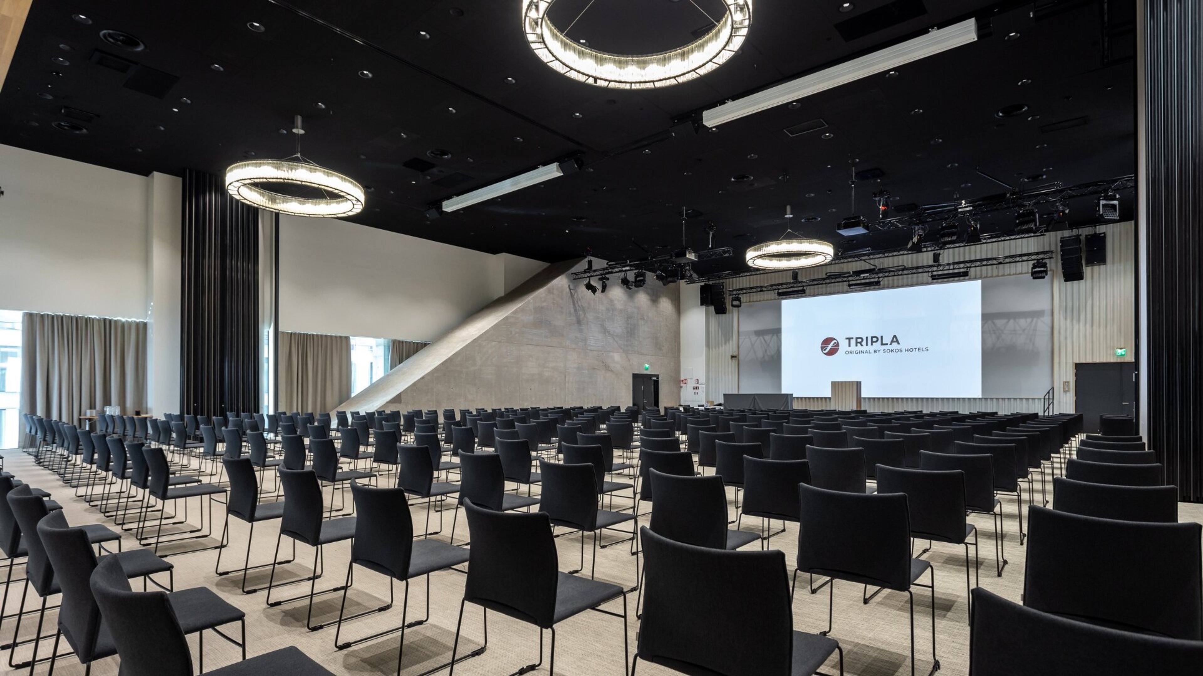 Moderni kokoustila, jossa rivit mustia tuoleja, valkoinen lattia ja korkea katto rengasmaisilla valaisimilla. Edessä valkokangas.