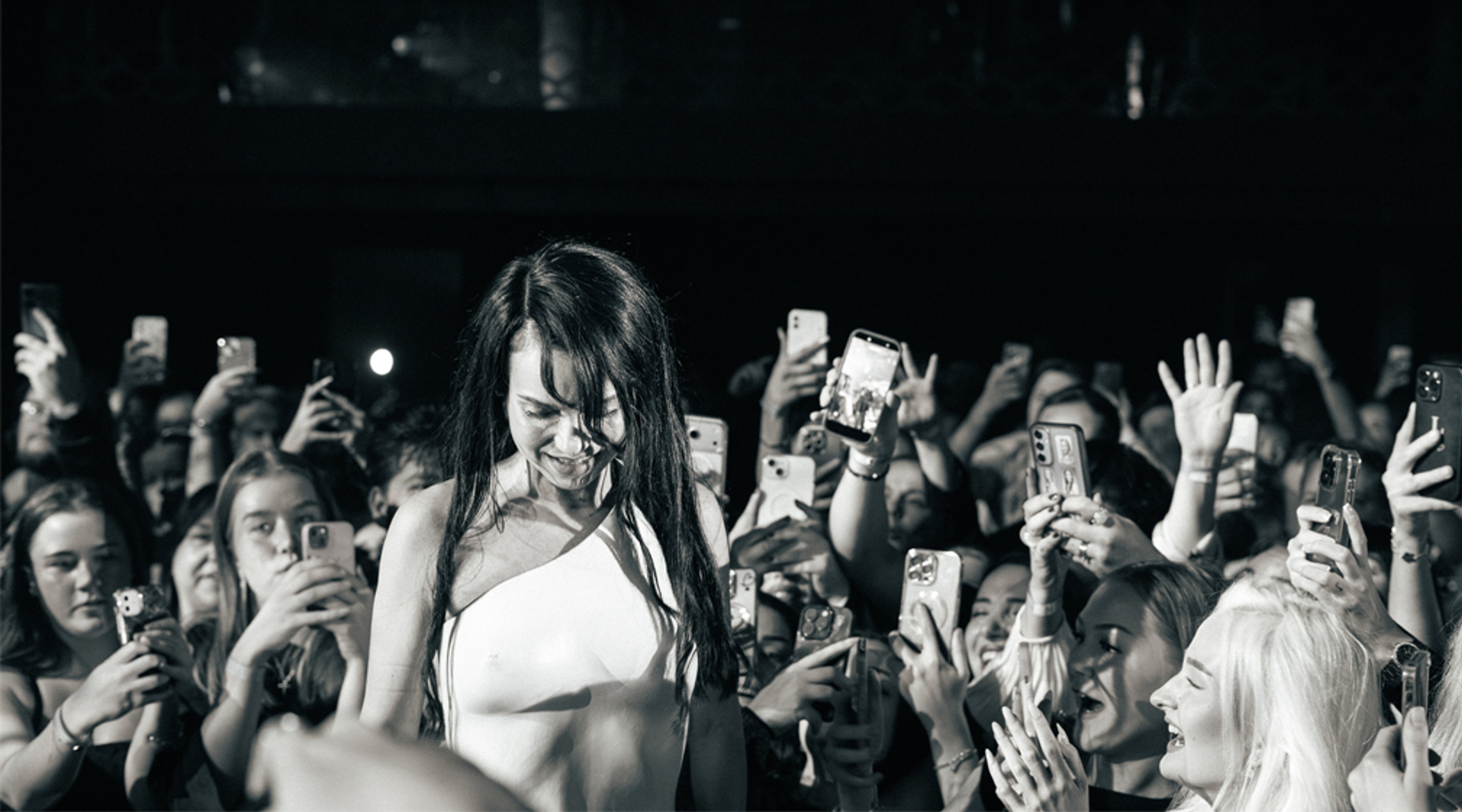 Henkilö valkoisessa vaatteessa esiintymislavalla, taustalla innokas yleisö nostaa kännyköitään ilmaan.