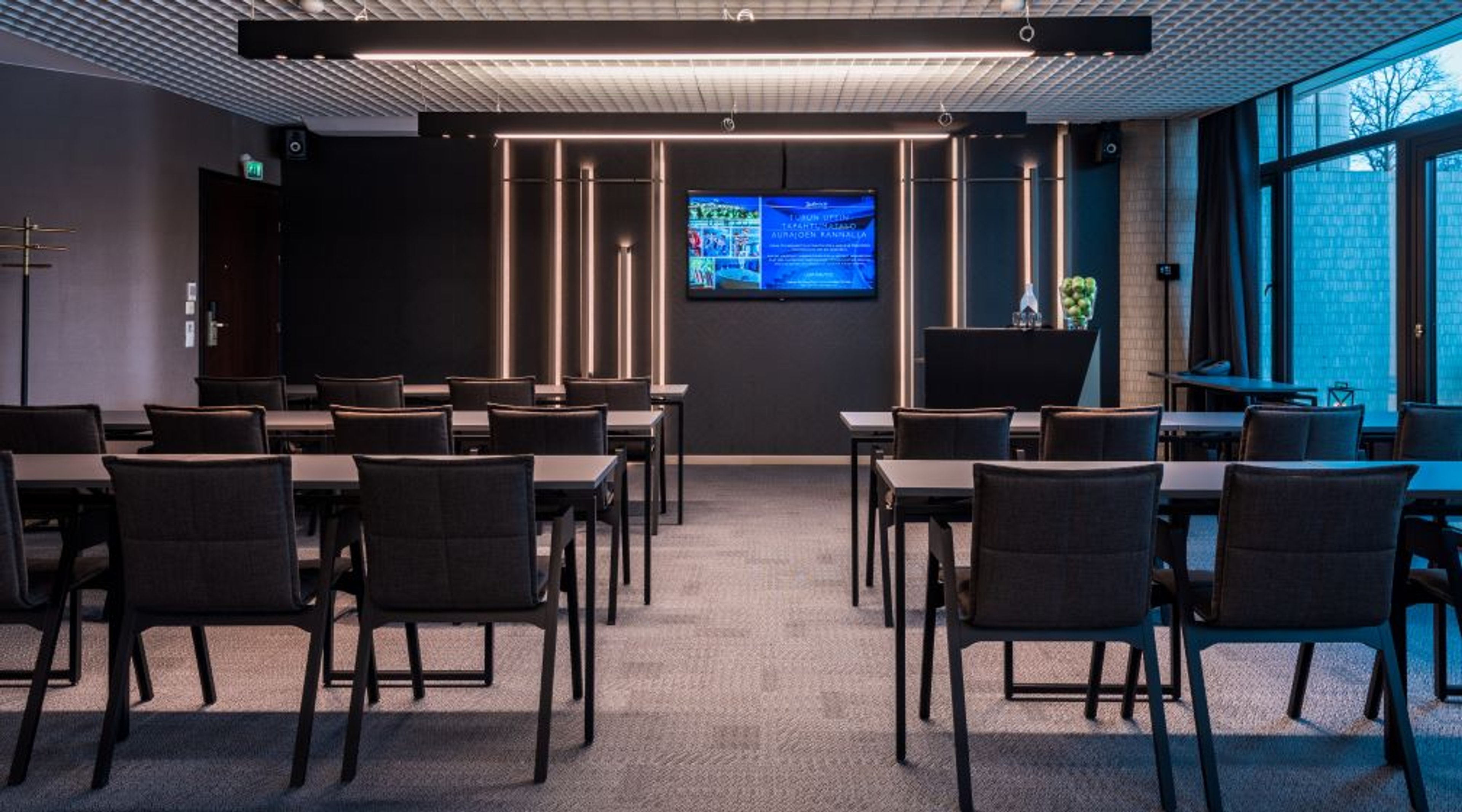 Modern conference room with rows of tables, dark chairs, wall-mounted screen and decorative light patterns. Level floor surface and wide passage space.