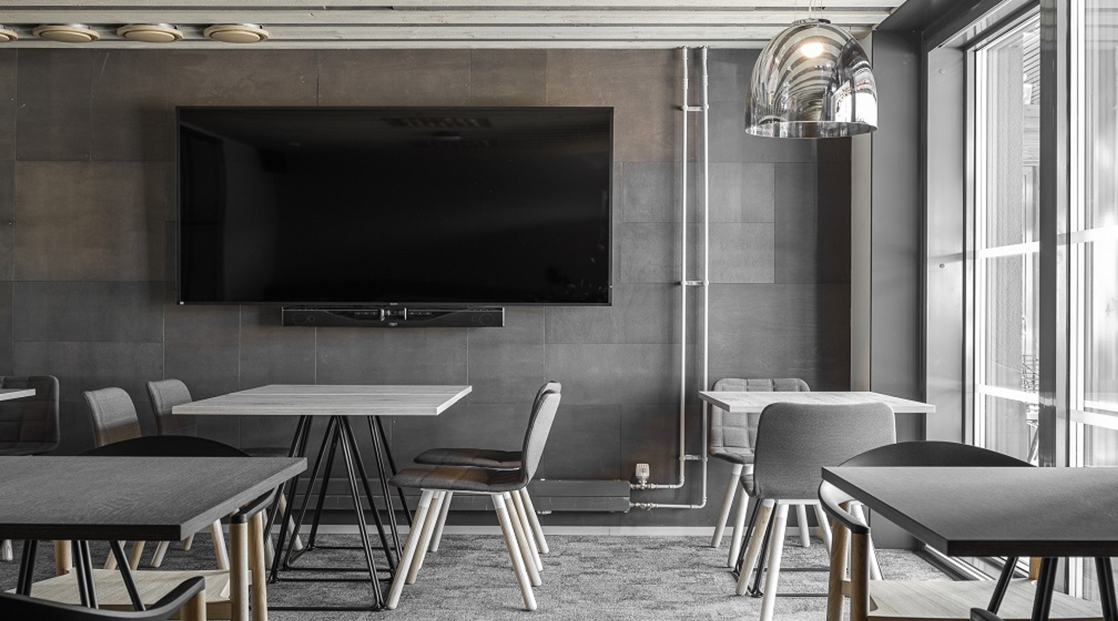 Moderni neuvottelutila, jossa harmaat pöydät ja tuolit, seinään kiinnitetty suuri televisio sekä isot ikkunat tuovat luonnonvaloa.