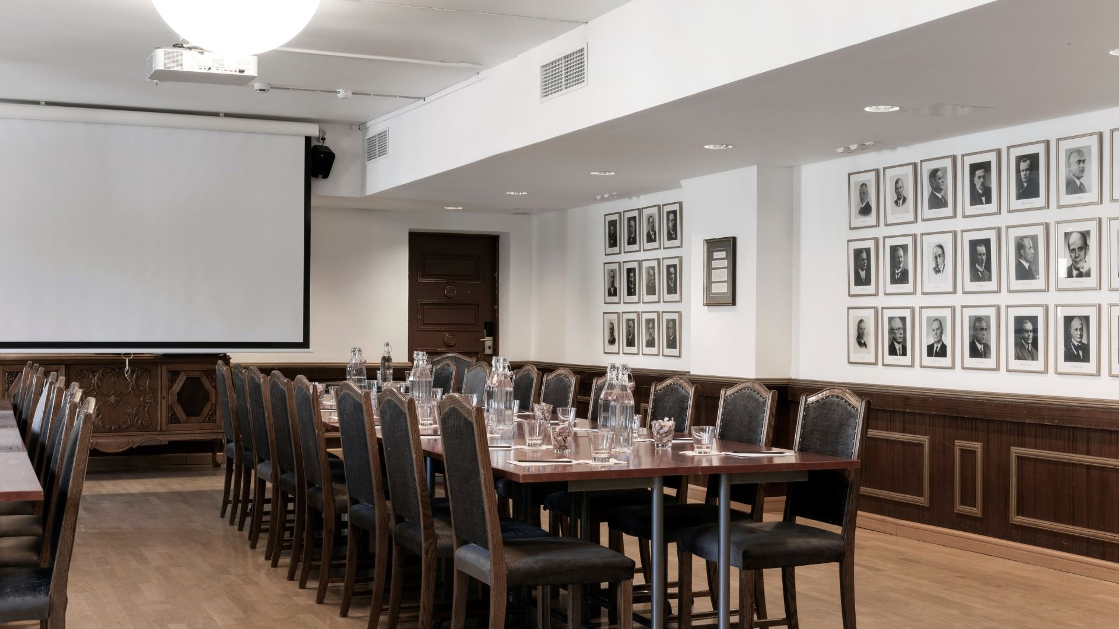 Kokoushuone, jossa on pitkä pöytä tuoleineen, projektori, valkokangas ja muotokuvia seinillä. Puulattia ja hyvä valaistus.