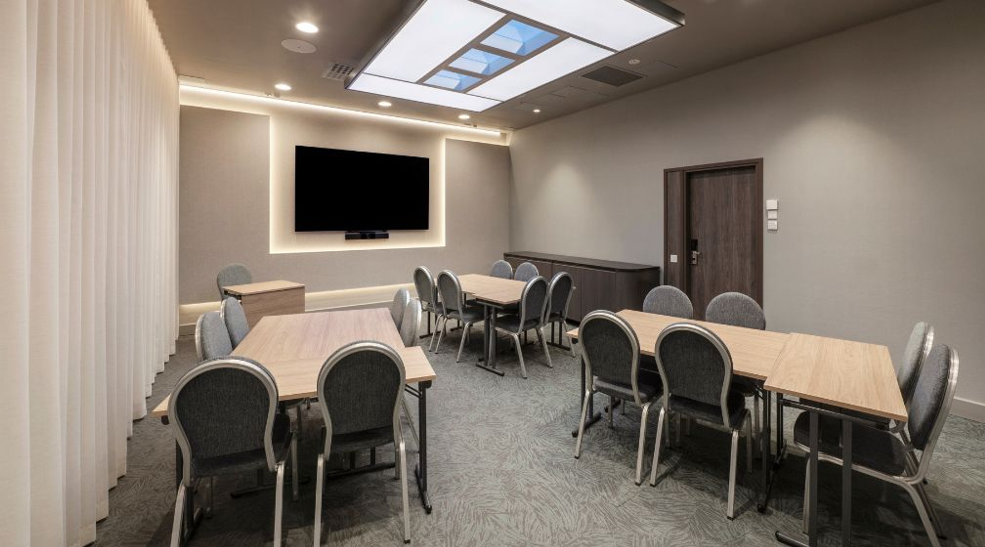 Moderni kokoushuone, jossa kolme puupintaista pöytää, harmaat tuolit, seinällä musta näyttö ja kattovalaistus kattoikkunalla.