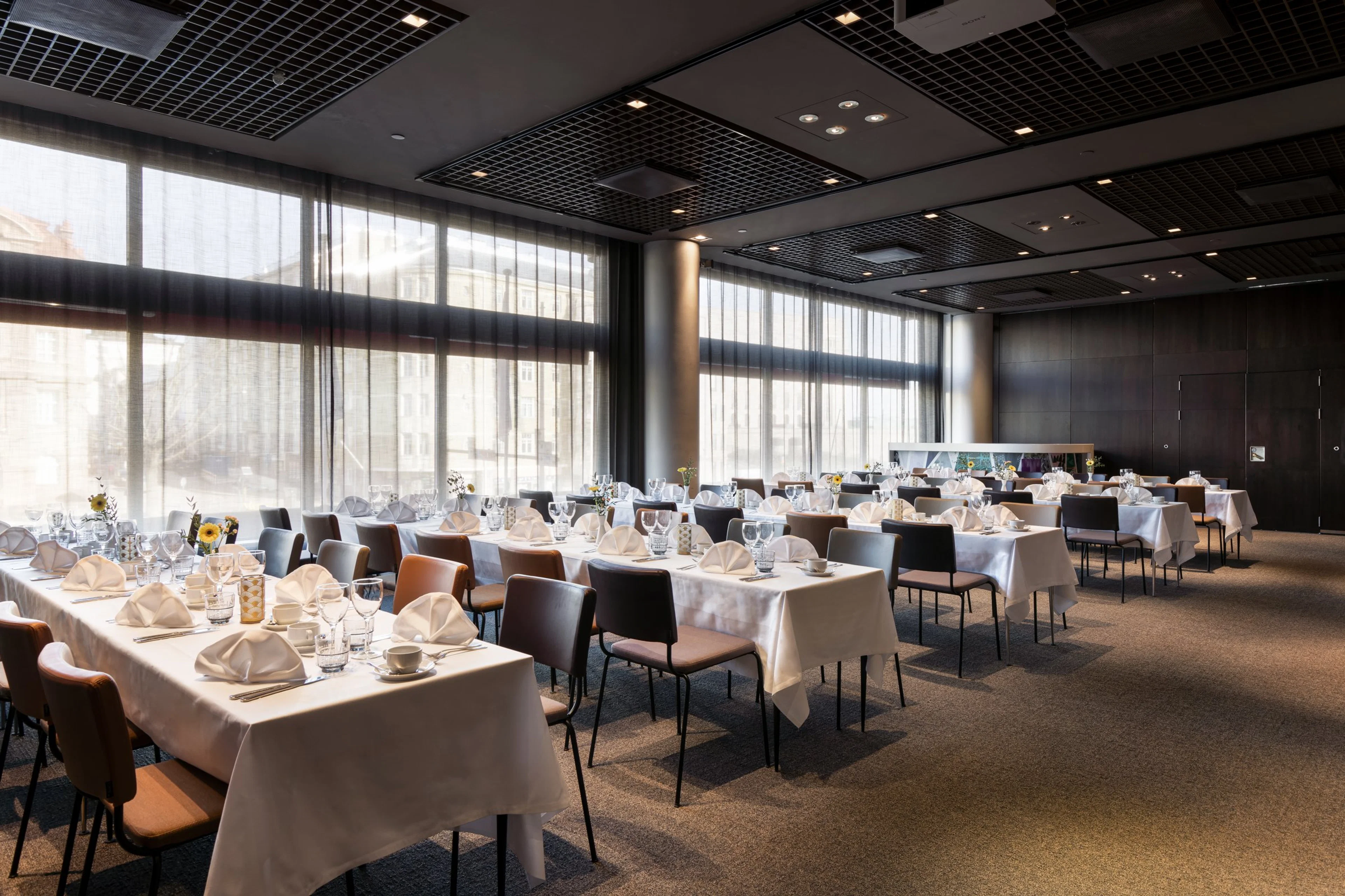 Ravintolasali, jossa on valkoliinaiset pöydät kattauksineen. Suuret ikkunat tuovat luonnonvaloa tilaan. Lattia on tasainen ja tilava.