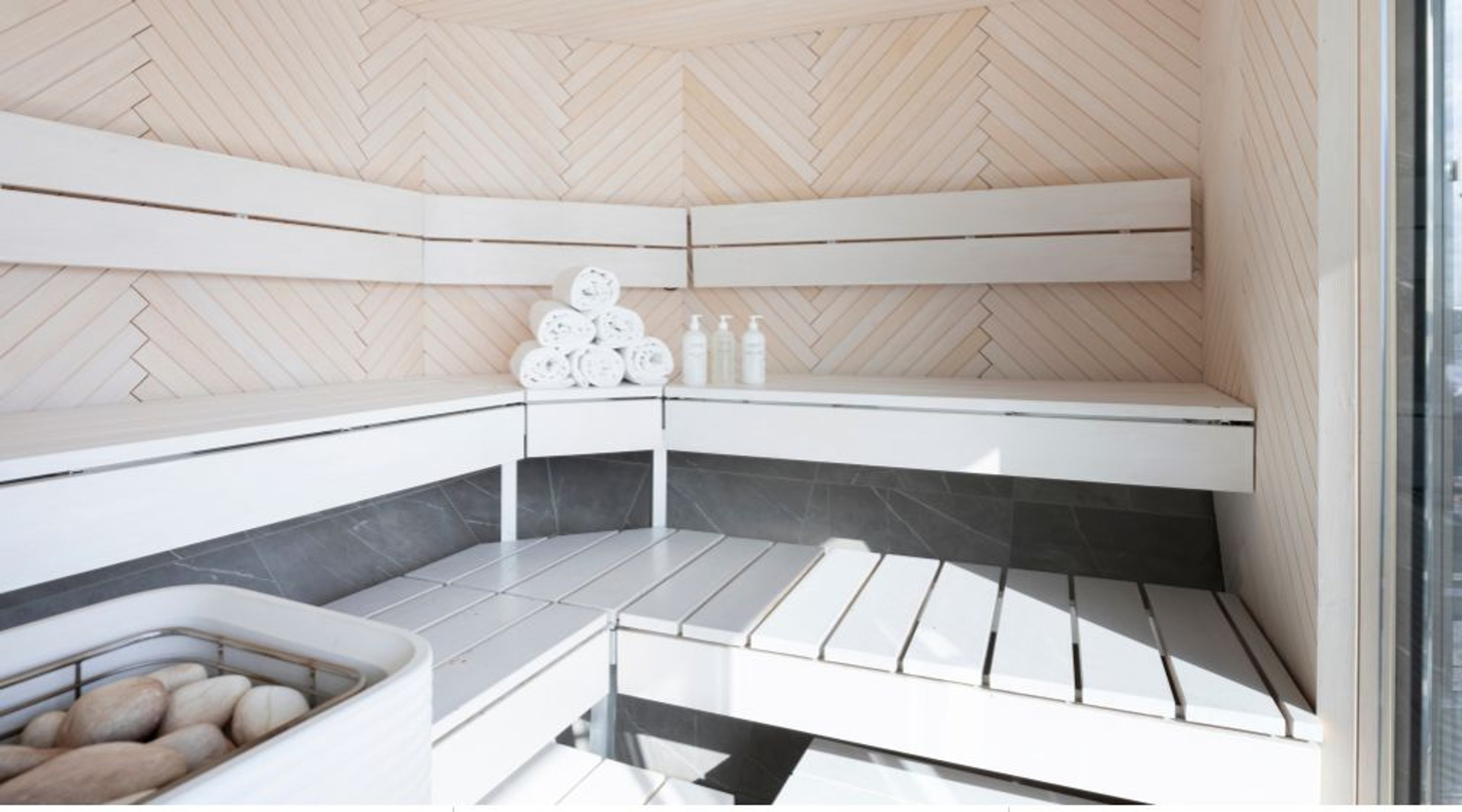 Moderni sauna, jossa vaaleaa puu kalanruotokuviossa. Saunan tasot ovat kaksikerroksiset ja valkoisia, lattia on tummanharmaa. Nurkalla on pyyhkeitä ja pulloja.