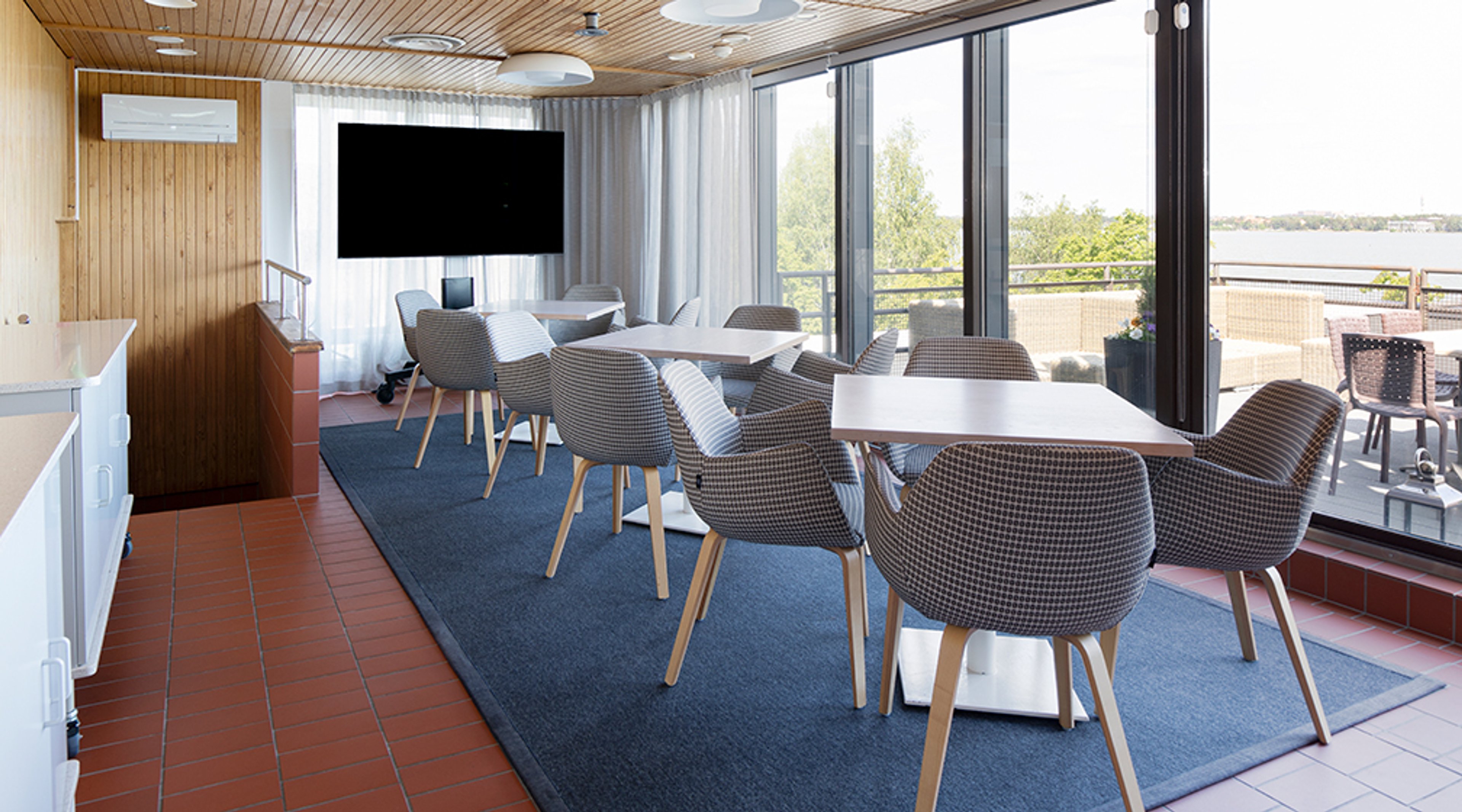 Moderni neuvotteluhuone puupaneeliseinillä, suurilla ikkunoilla ja näkymällä ulos. Tilassa on valkoisia pöytiä ja kuviollisia tuoleja sinisen maton päällä.