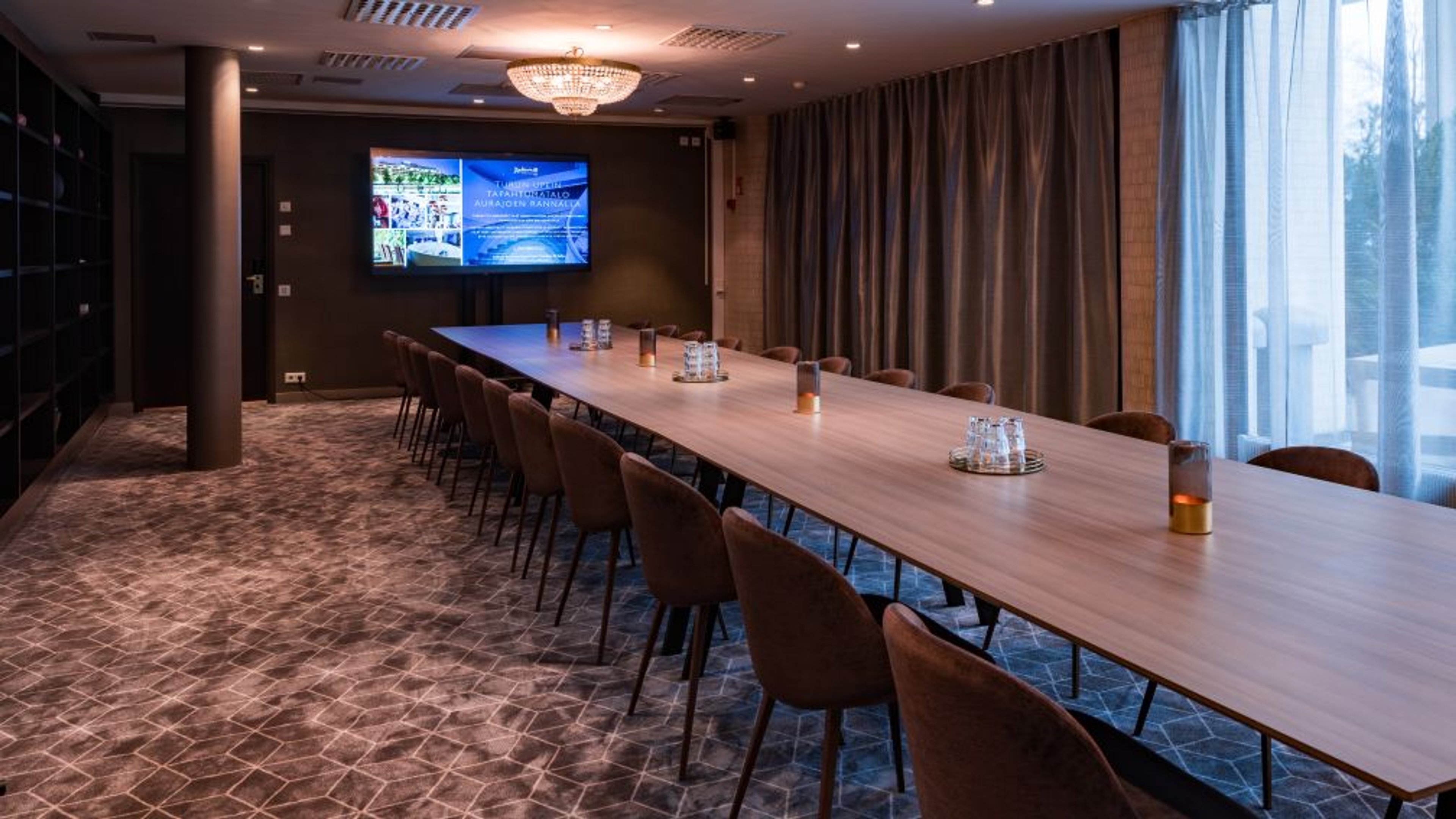 Moderni kokoustila pitkällä puupöydällä ja ruskeilla tuoleilla. Taustalla näyttö, kristallikruunu katossa ja suuret ikkunat luonnonvalolla.