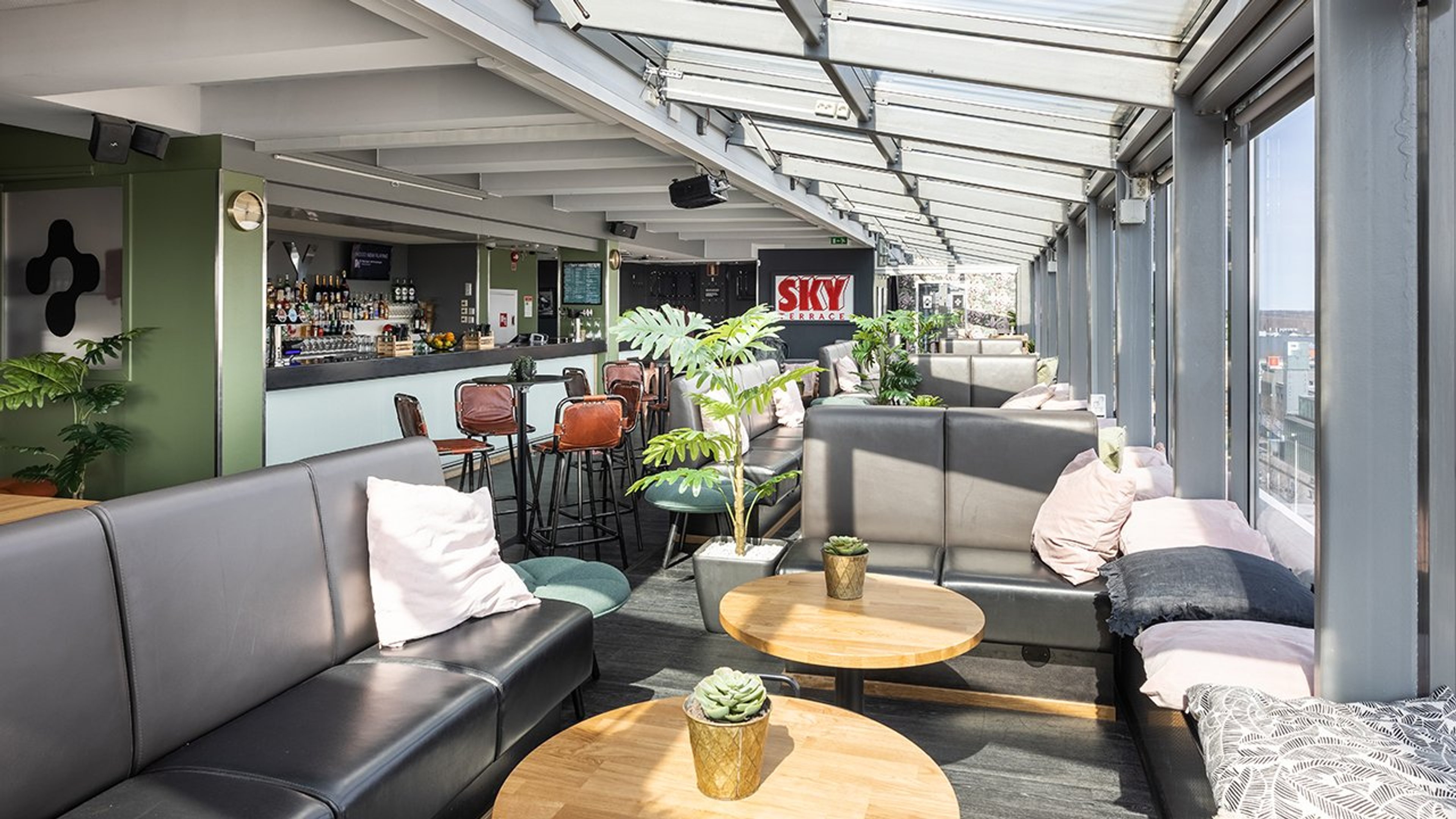 Valoisa kattoterassibaari, jossa harmaita sohvia, puisia pöytiä ja viherkasveja. Tilava kulkutila ja lasikatto tuovat luonnonvaloa.