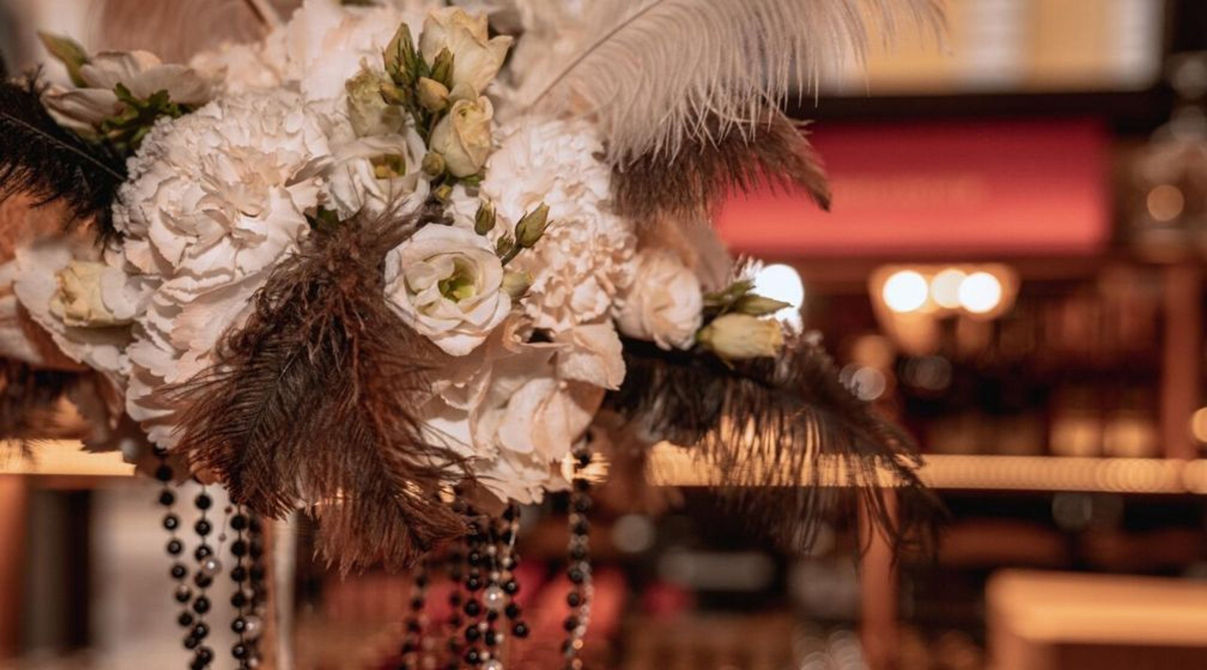 Koristeellinen kukka-asetelma valkoisista ruusuista ja neilikasta sulkien ja mustien helmien kanssa, hämyisässä juhlatilassa.