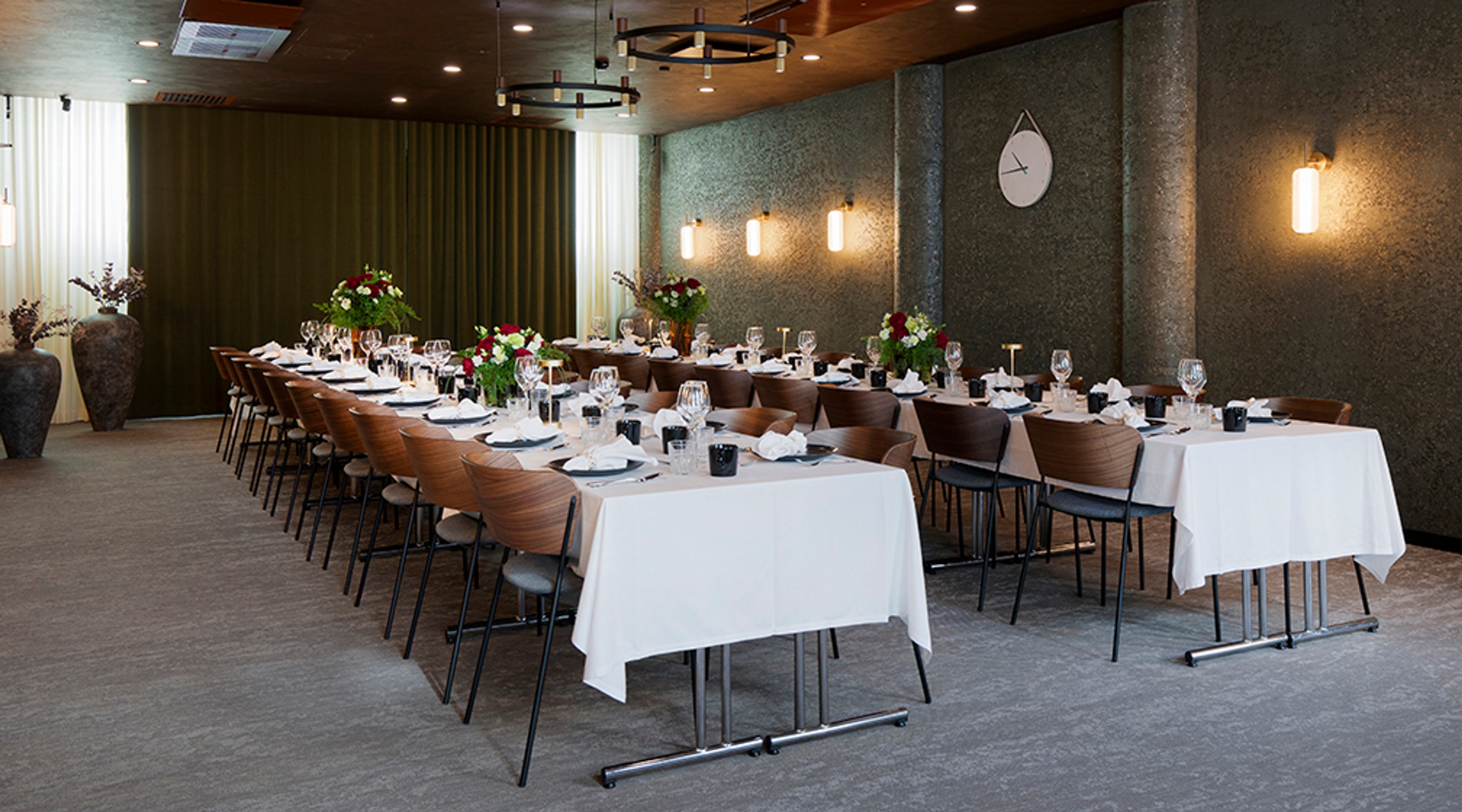 Tyylikäs ravintolan yksityistila pitkällä pöytäjärjestelyllä, valkoiset pöytäliinat ja kukkakoristeita. Seinissä lämpimät valaisimet ja takaseinällä kello.