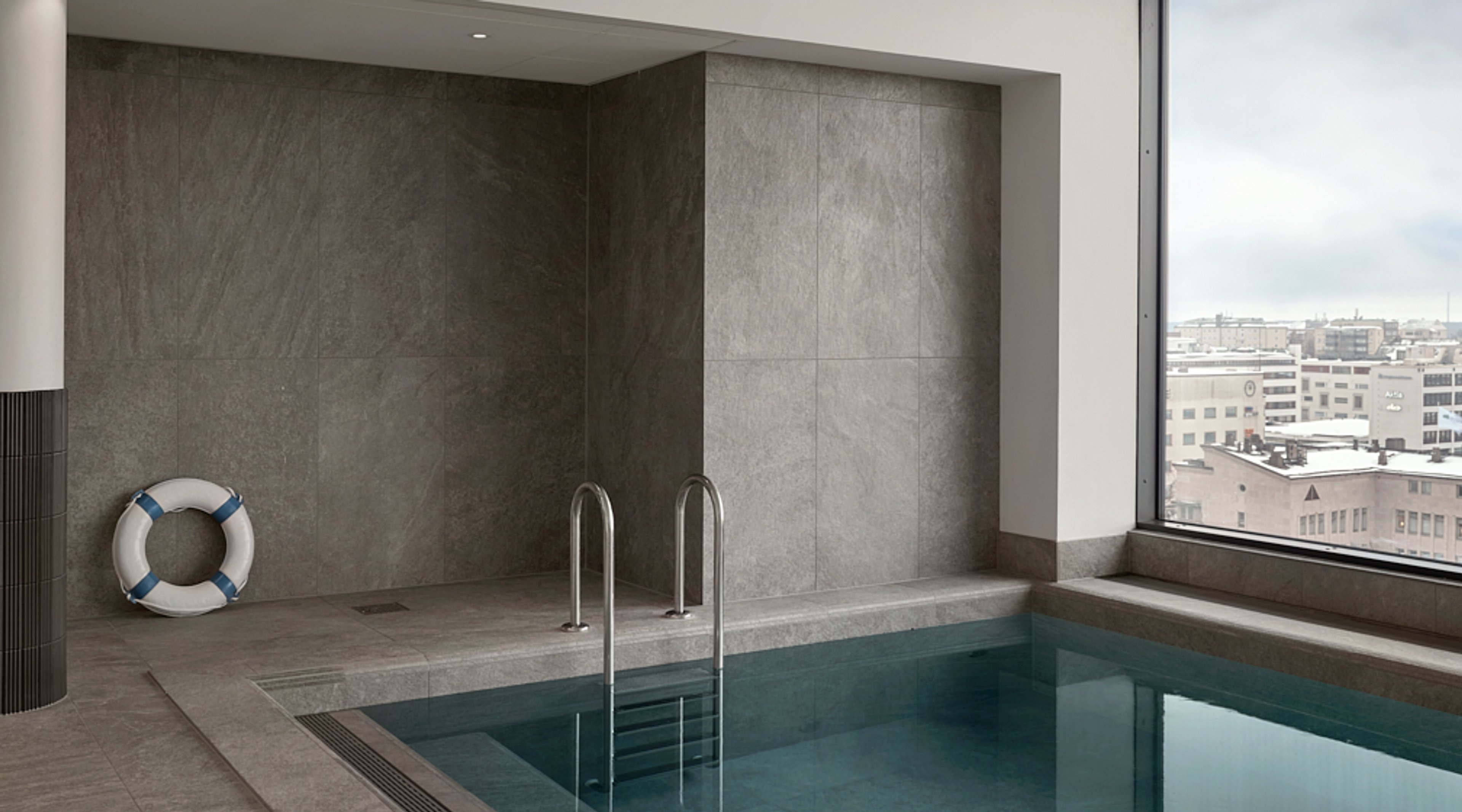 Sisäuima-allas harmailla kivilaatoilla vuoratulla alueella. Metallinen uimatikkaat altaaseen, pelastusrengas seinällä ja suuresta ikkunasta kaupunkinäkymä.
