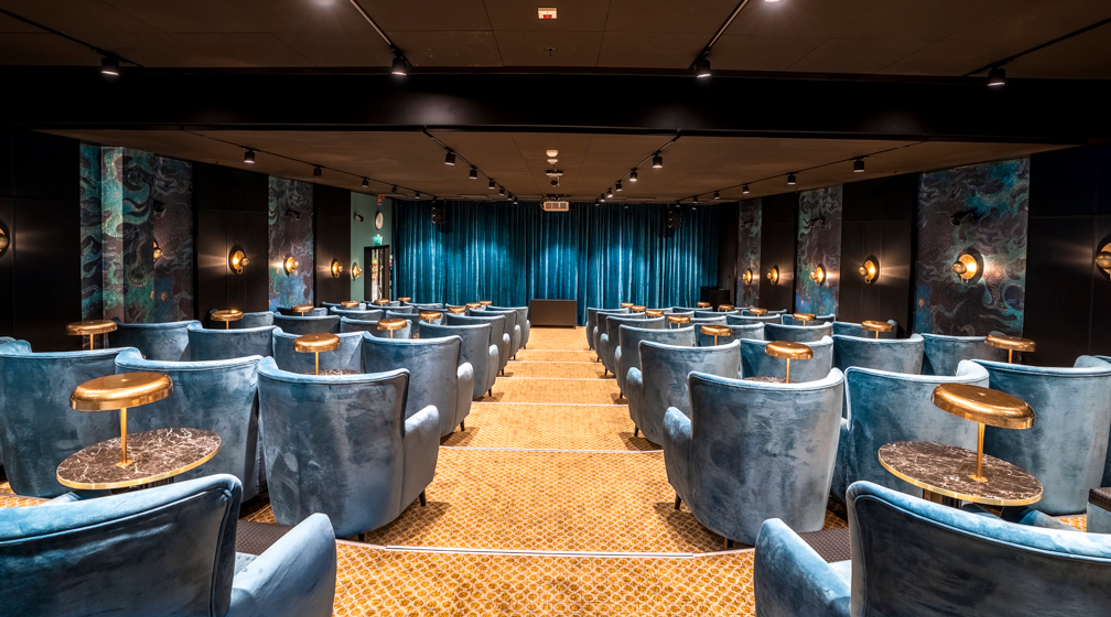 Moderni auditorio turkooseilla samettinojatuoleilla ja pienillä marmoripöydillä. Turkoosin verhon edessä on esiintymistila ja tilassa on kuviollinen oranssi matto.