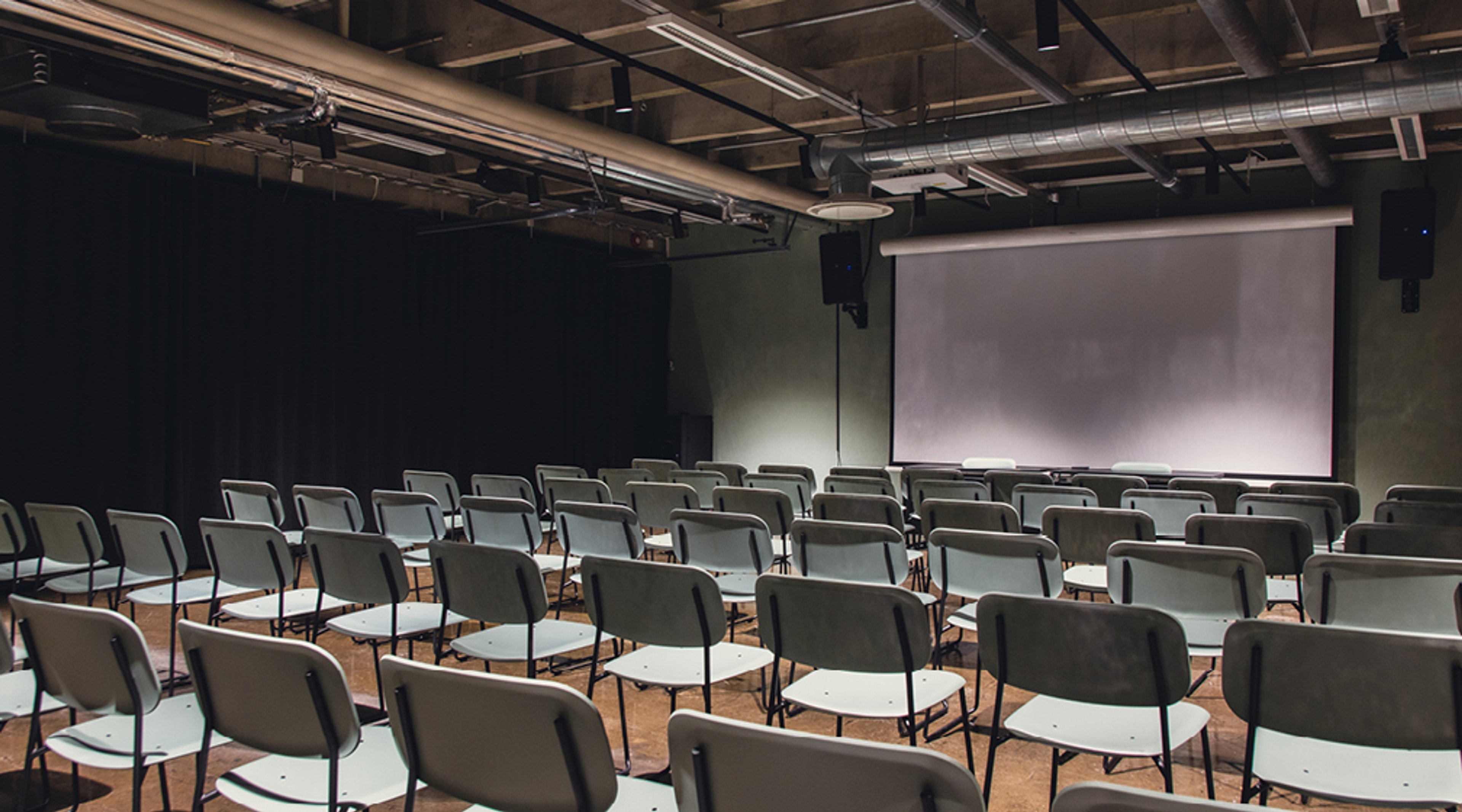 Kokoustila, jossa on useita tuolirivistöjä teatterimuodossa. Edessä valkoinen esityskangas ja tumma sisustus.