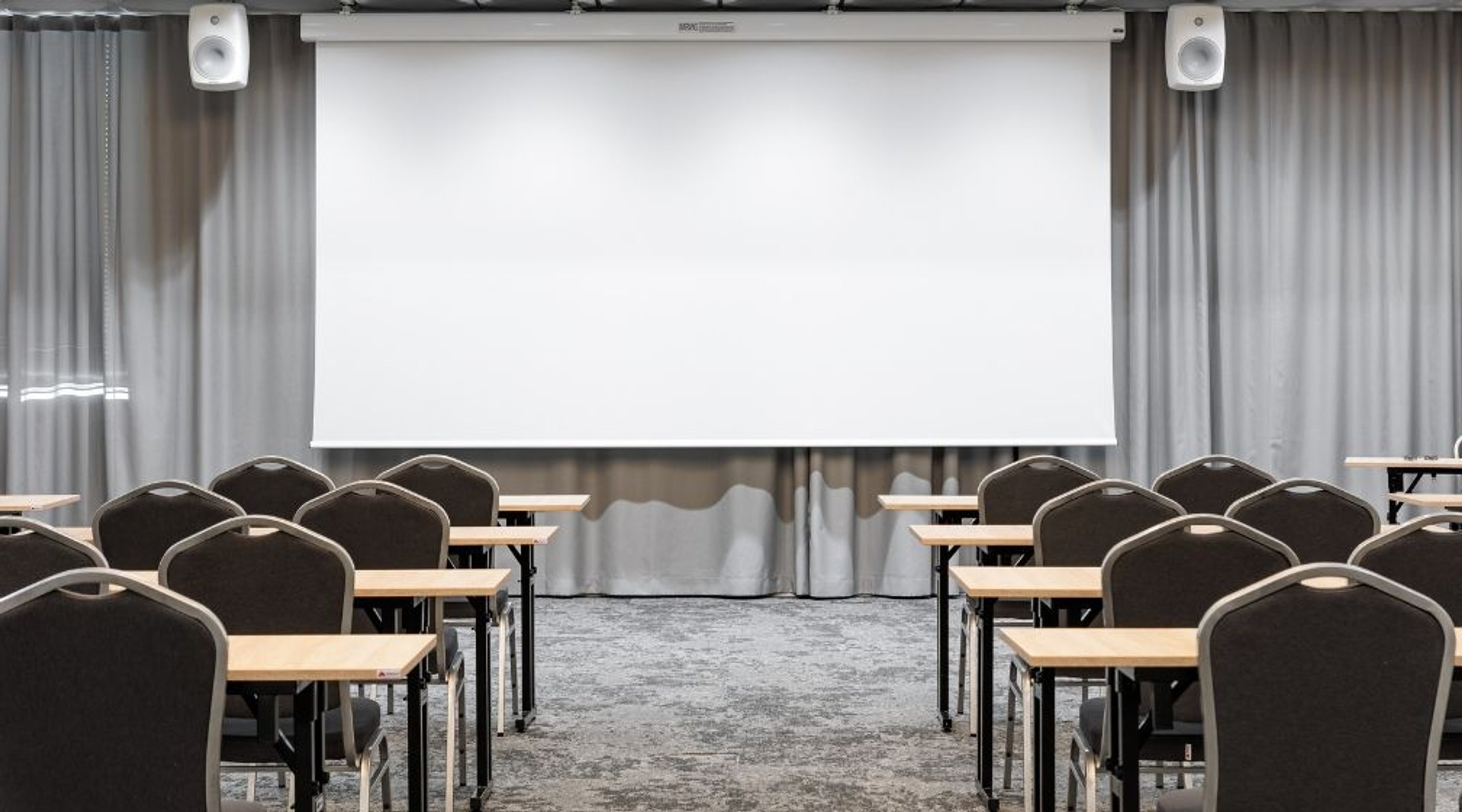 Konferenssihuone, jossa pöydät ja tuolit on aseteltu riveihin projektorikankaan eteen. Harmaa kokolattiamatto ja vaaleanharmaat verhot.