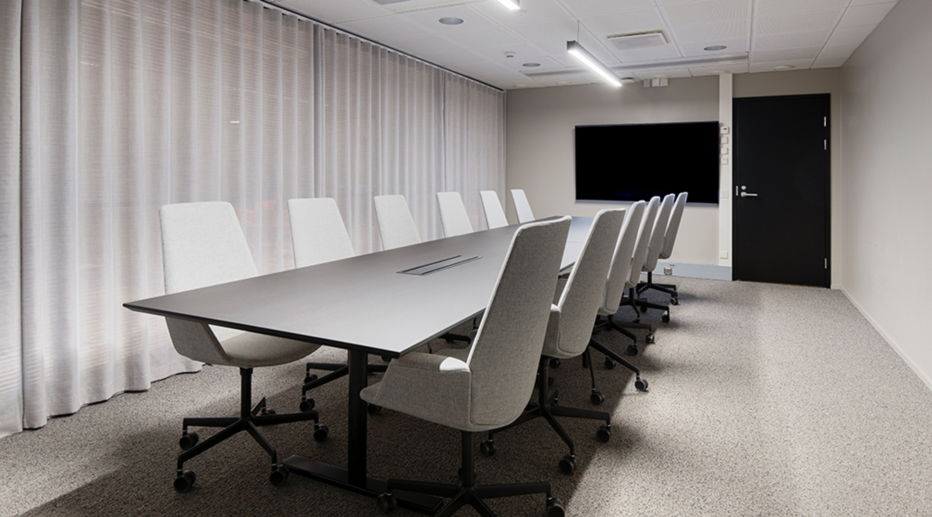 Modern conference room with a long table, white chairs, white curtain and a screen on the wall. The room has hard flooring and a wide door.