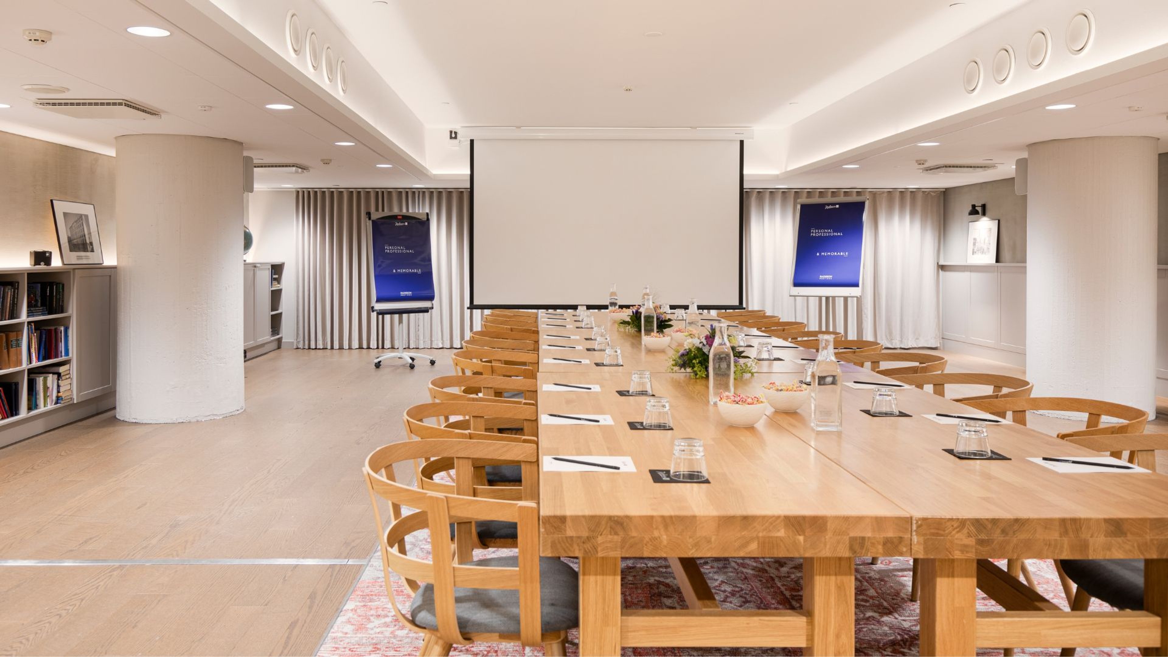 Moderni kokoustila, jossa pitkä puinen pöytä puisilla tuoleilla, valkoinen projektorikangas, tasainen puulattia ja hyvä valaistus katossa.