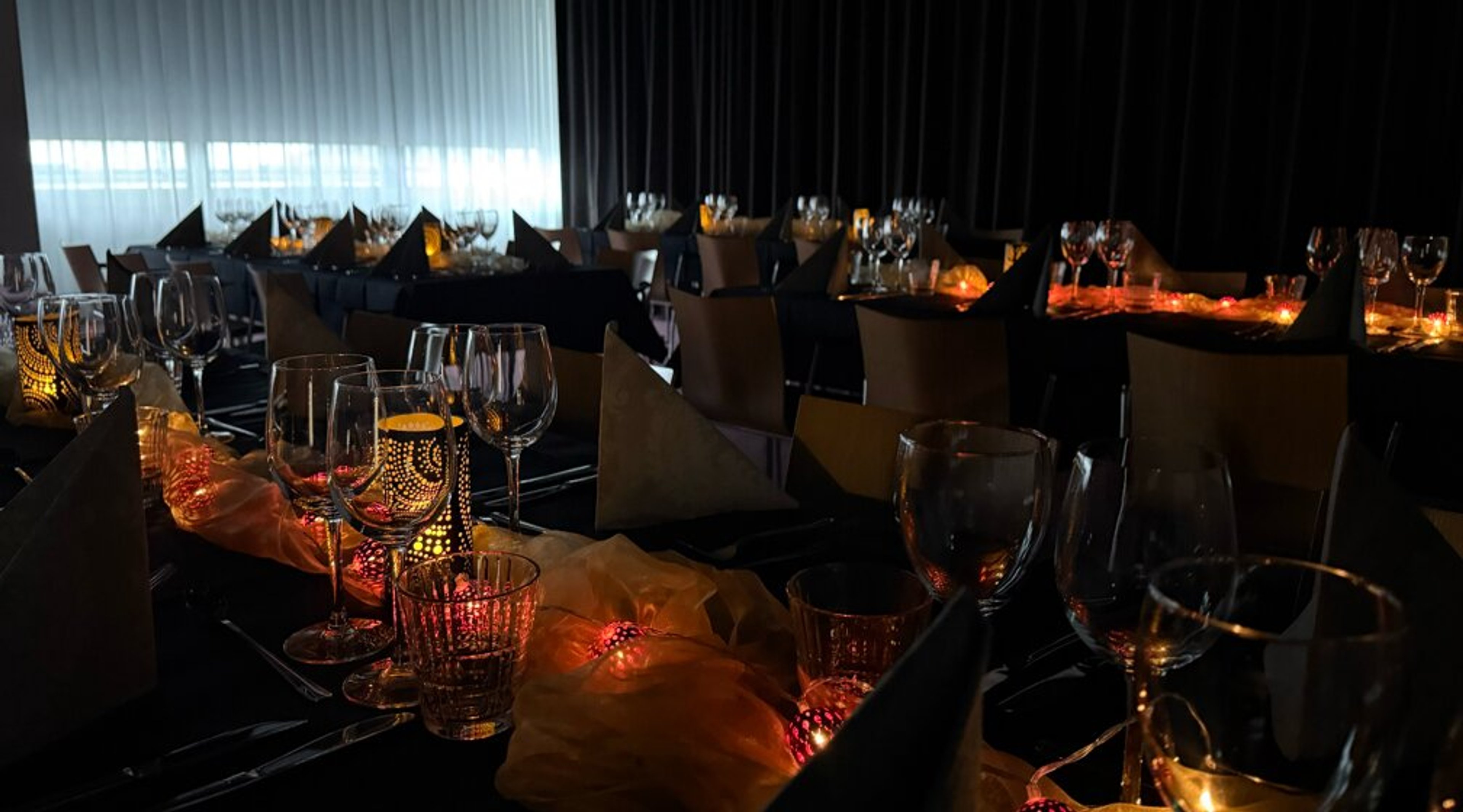 Hämaras valgustatud söögisaal laudadega reas. Lauad on kaunistatud veiniklaasidega ja dekoratiivsete valgustatud laternatega. Toolid on tumedad.